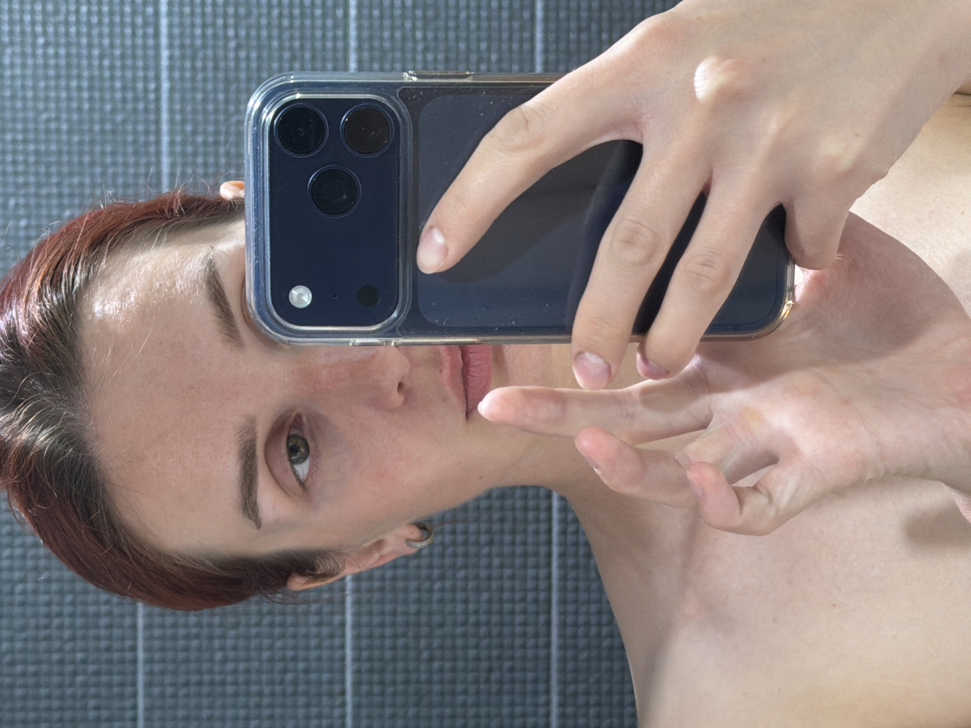 A person is holding a mobile phone in front of their face, seemingly taking a selfie. The phone case is transparent with a dark-colored phone. The person has light skin and auburn hair. The background is a gray textured wall. The location, Chaikovsky, Russia, is based solely on the user's prompt, there is no other indication of location in the image.