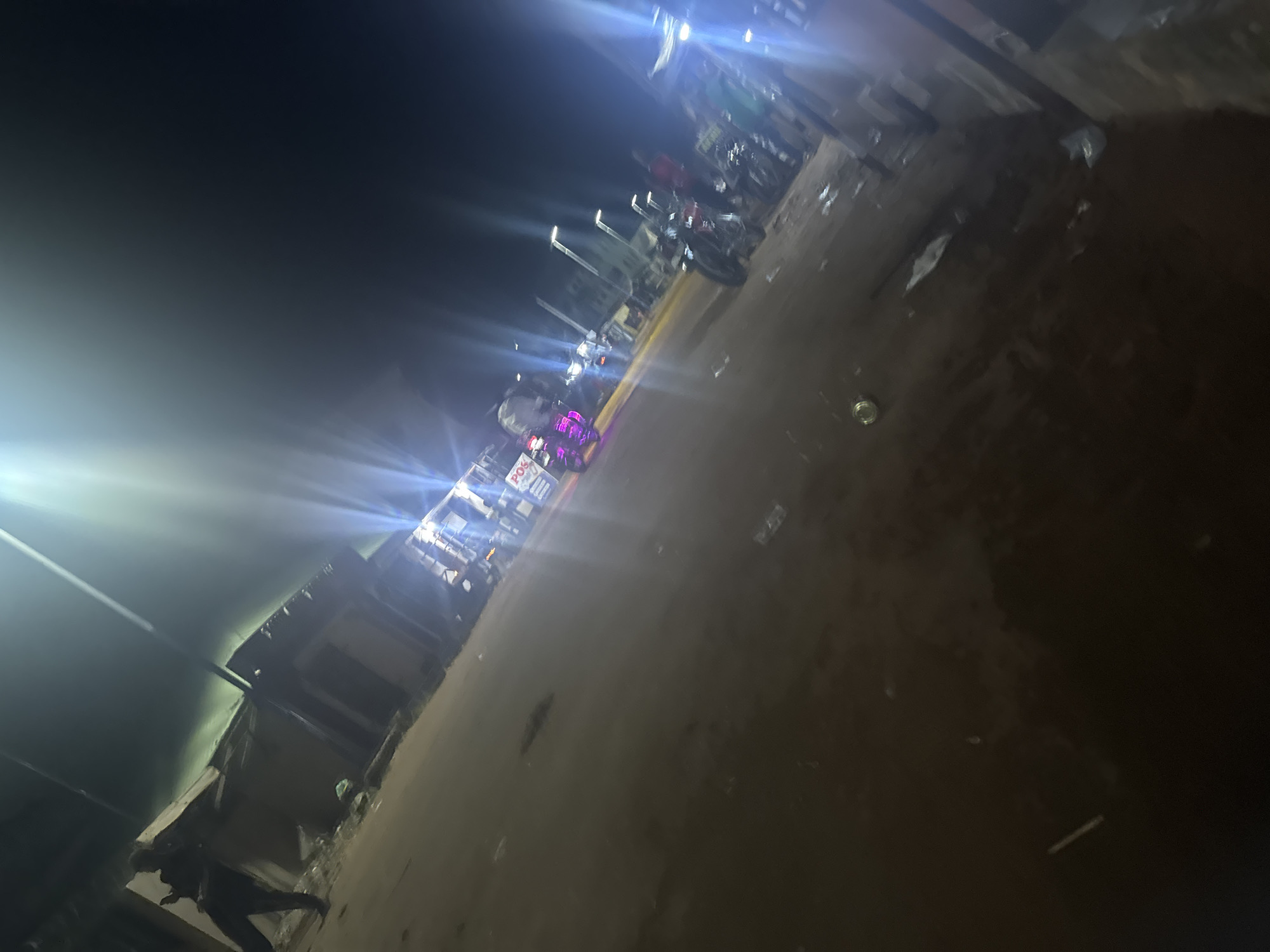 The image shows a street scene in Ihiagwa, Nigeria at night. Streetlights and lights from businesses illuminate the scene. There are parked motorcycles along the side of the street and people standing in groups. Buildings line the street, some appearing to be shops or businesses. The ground is uneven and appears to be a mix of dirt and pavement. There is trash scattered on the ground.