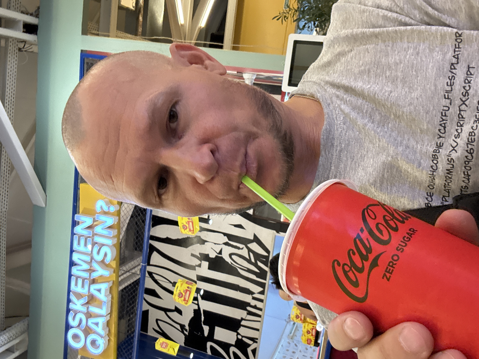 A close-up, selfie-style image shows a bald, fair-skinned man with a goatee drinking from a red Coca-Cola Zero Sugar cup with a green straw. He is wearing a grey t-shirt with black text printed on it. In the background, a sign with bright blue, illuminated letters reads "OSKEMEN QALAYSIN?". This phrase is Kazakh for "How are you, Oskemen?". The sign is mounted above a black and white graphic pattern. Yellow decorative items are hanging from the ceiling. The setting appears to be an indoor public space, possibly a cafe or entertainment venue, in Oskemen, Kazakhstan.