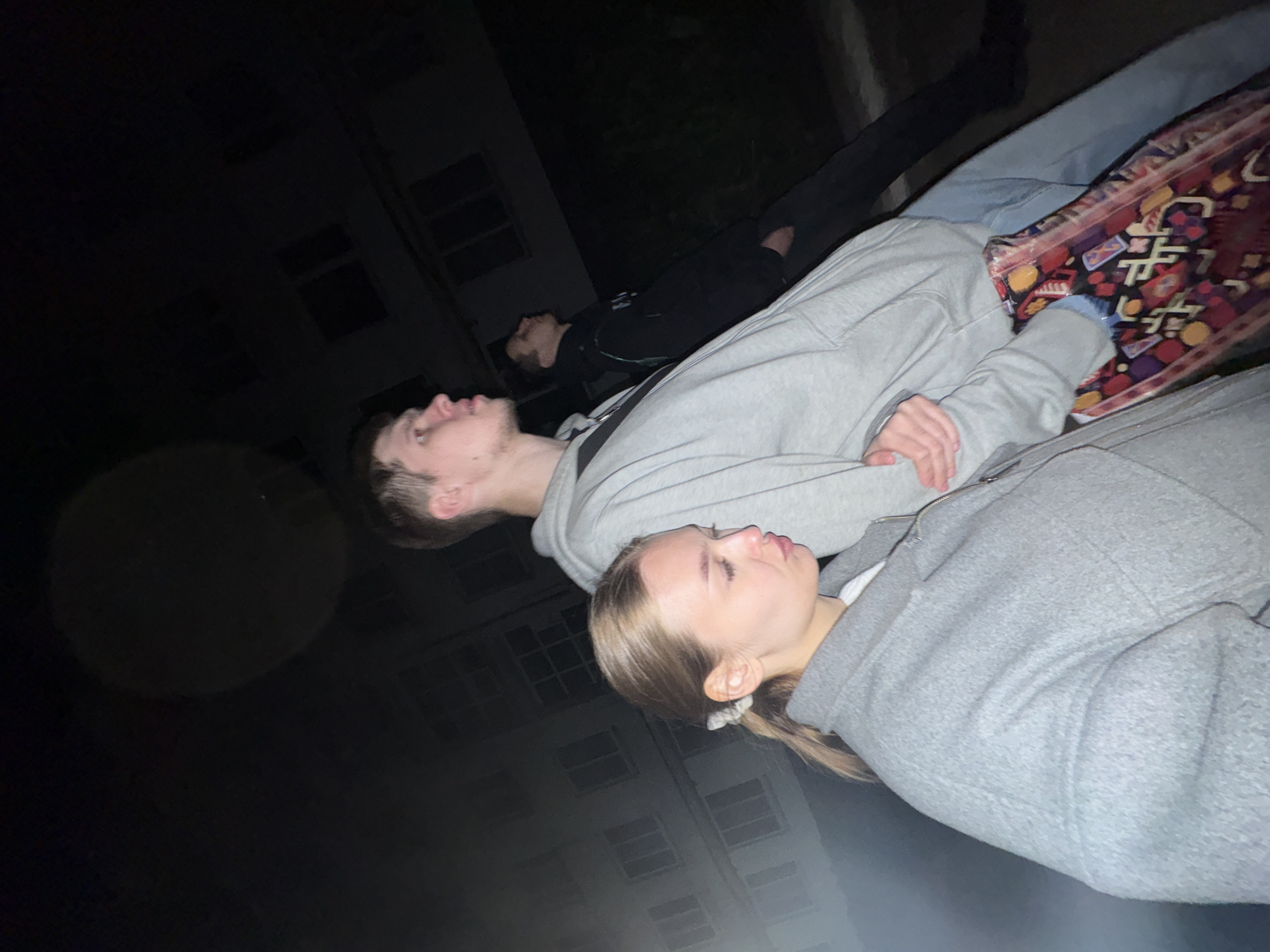 Three people are present outdoors at night in Chaikovsky, Russia. The backdrop features a multi-story building with visible windows. A woman in the foreground wears a gray zippered jacket, while a man behind her wears a gray hoodie. Further behind them, another man is dressed in dark clothing and jeans. A patterned bag rests beside the man in the hoodie. All three are looking upwards.