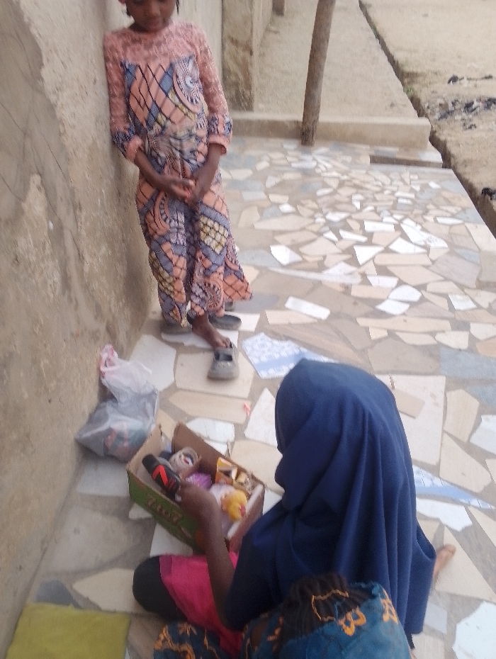 A girl in an orange and blue dress leans against a wall next to a mosaic tiled patio. Another girl wearing a blue hijab sits on the patio floor with a shoebox filled with colorful objects. A plastic bag sits nearby. The girls are outside near a tree, and the patio extends into a path near bare earth.