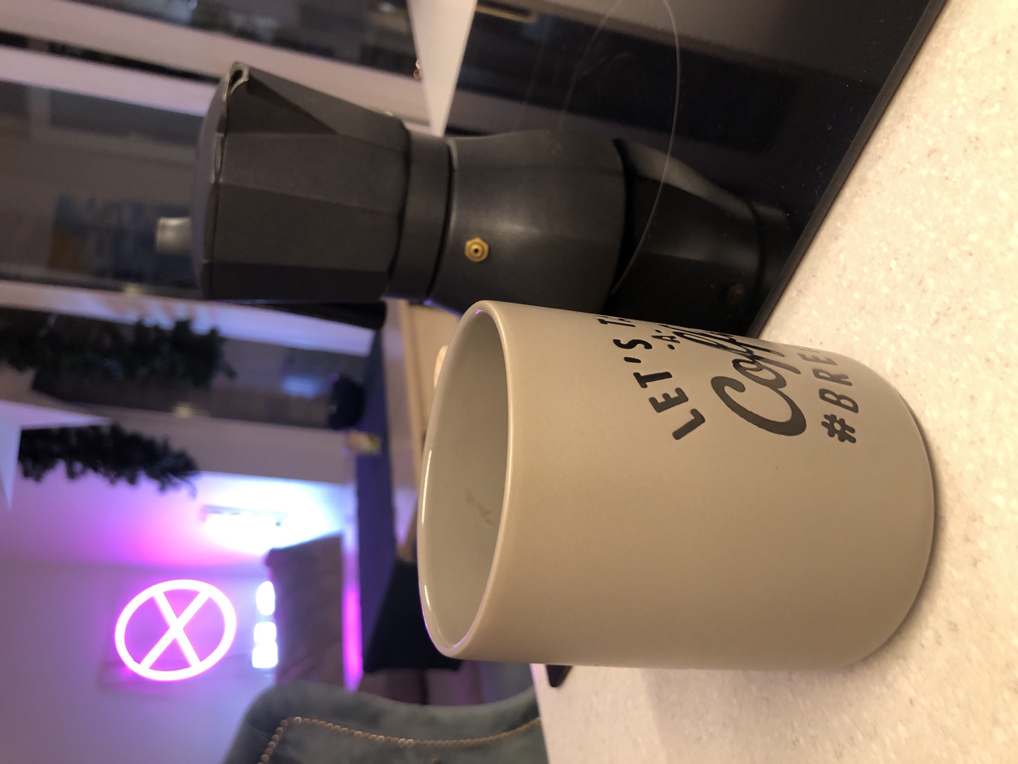 A still life image captures a kitchen counter scene with coffee-making items in Saint Petersburg, Russia. A black, angular Moka pot with a brass pressure release valve rests on a sleek black induction hob, positioned towards the top left of the frame. Adjacent to it, on a light-colored, speckled countertop, sits an empty light beige cylindrical coffee mug. Black text on the mug's side reads "LET'S COFFEE" with a partially visible hashtag, likely "#BREW."
The background, largely out of focus, reveals an indoor setting with artificial lighting. A prominent neon sign, glowing pink and shaped like a circle with an 'X' or cross inside, adds a vibrant pop of color to the blurred backdrop. Further back, dark green foliage, possibly a garland or decoration, is vaguely discernible, along with hints of furniture like a chair with stud detailing. The overall lighting suggests an interior shot, possibly in the evening or early morning, with ambient and artificial light sources. The arrangement suggests a moment before or after enjoying coffee, highlighting a daily ritual.