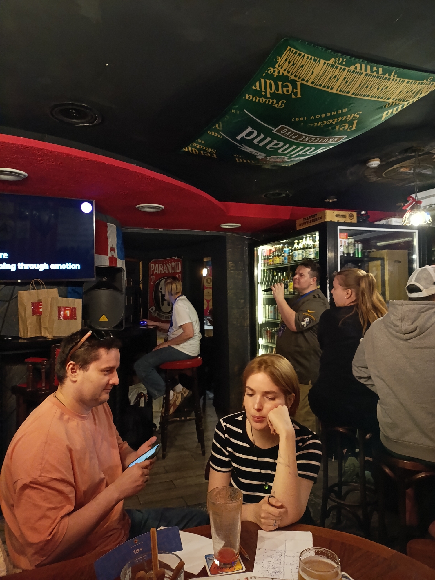 In an indoor establishment, potentially a bar or pub, a group of individuals are seen. A man in an orange t-shirt is seated, looking at his phone. Beside him, a woman with blonde hair and a black and white striped shirt rests her head on her hand, appearing pensive. In the background, a man in a green shirt holds a microphone and speaks into it, facing a wall of refrigerators stocked with beverages. Another woman with blonde hair tied in a ponytail is standing next to him. The ceiling is adorned with a large green banner featuring yellow text. The environment is dimly lit, with red accents on the ceiling and bar area. The overall impression is a casual gathering or event.