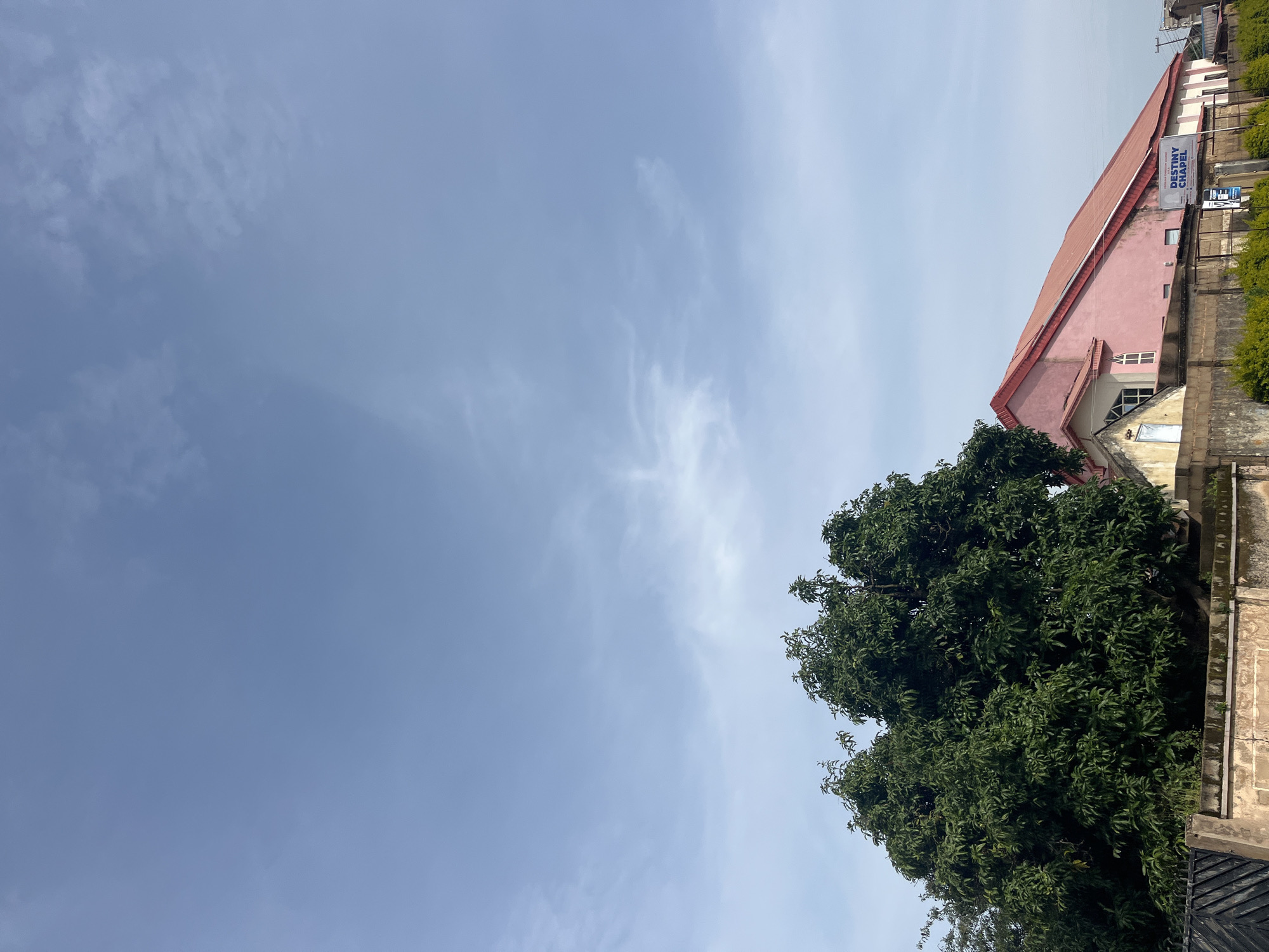 The image depicts the exterior of a building identified as "DESTINY CHAPEL" in Jos, Nigeria. The building has a prominent pink-colored, sloped roof and a lighter colored facade. To the left of the chapel, a large, dense green tree is visible. In the foreground, a concrete wall with some vegetation growing on top separates the street from the property. The sky above is mostly clear with some scattered wispy clouds. The overall scene suggests a daytime view of a religious building in an urban or semi-urban setting in Jos.