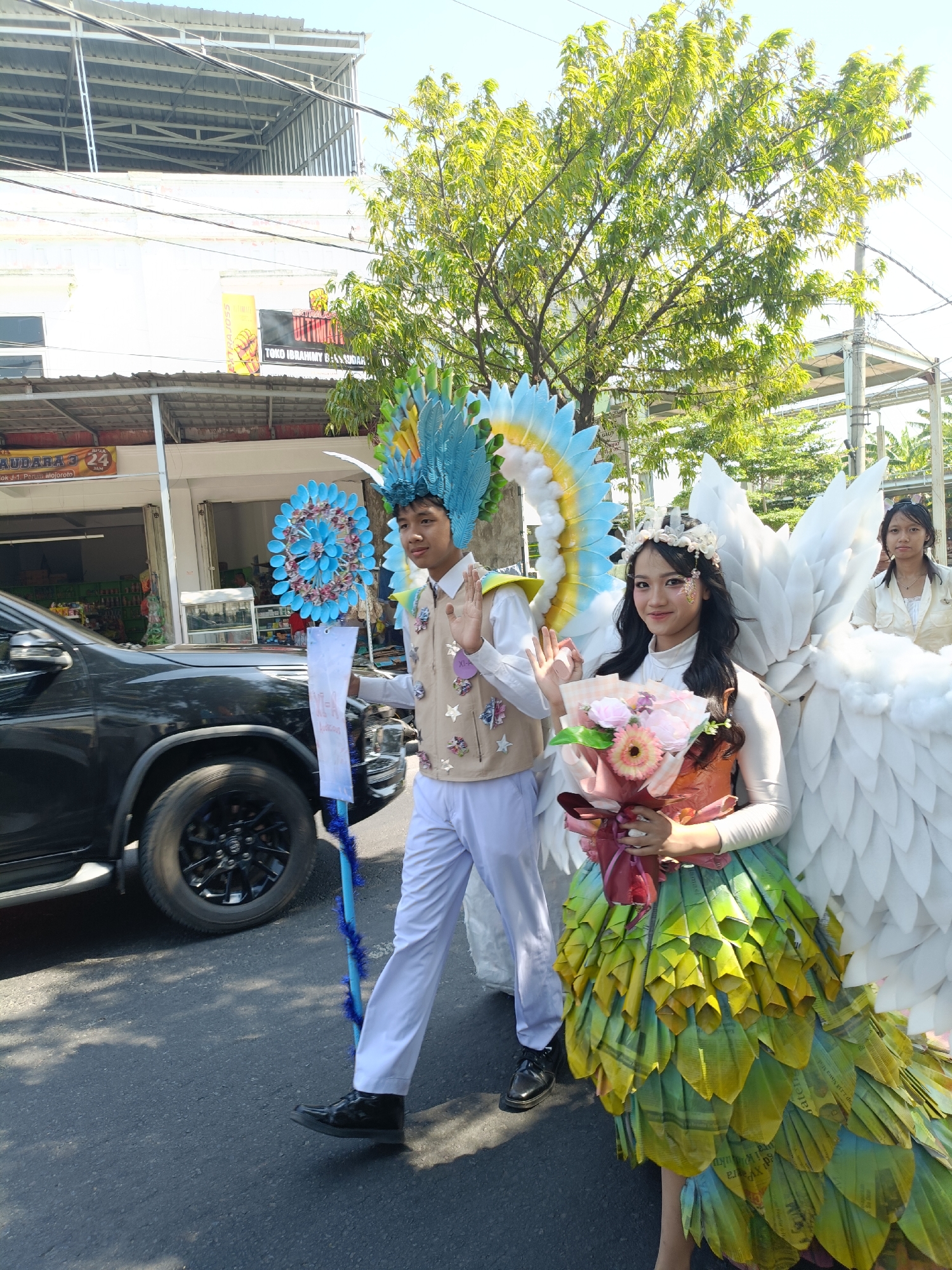 The image captures a street scene in Kediri City, Indonesia, possibly during a parade or cultural event. In the foreground, two individuals are dressed in elaborate costumes. The person on the left is a young male wearing a tall, blue and yellow feathered headdress, a light brown vest adorned with star-shaped embellishments, and white pants. He holds a blue staff with a circular blue decoration. The person on the right is a young female with a floral crown, a white long-sleeved top, and a striking skirt made of layered green and yellow leaf-like material. She has large white wings attached to her back and is holding a bouquet of pink and orange flowers. She is waving to the camera.
Behind them, a black SUV is partially visible on the left side of the frame. Further back, a building with a metal roof and signage is present. The signage includes text such as "PUTRAJOSS" and "TOKO IBRAHIMY GASSUDAR". In the background, trees with green foliage and a bright sky suggest a sunny day. Another young female in a light-colored outfit can be seen in the background to the right. The scene is set on an asphalt road, with some shadows cast on the ground.
