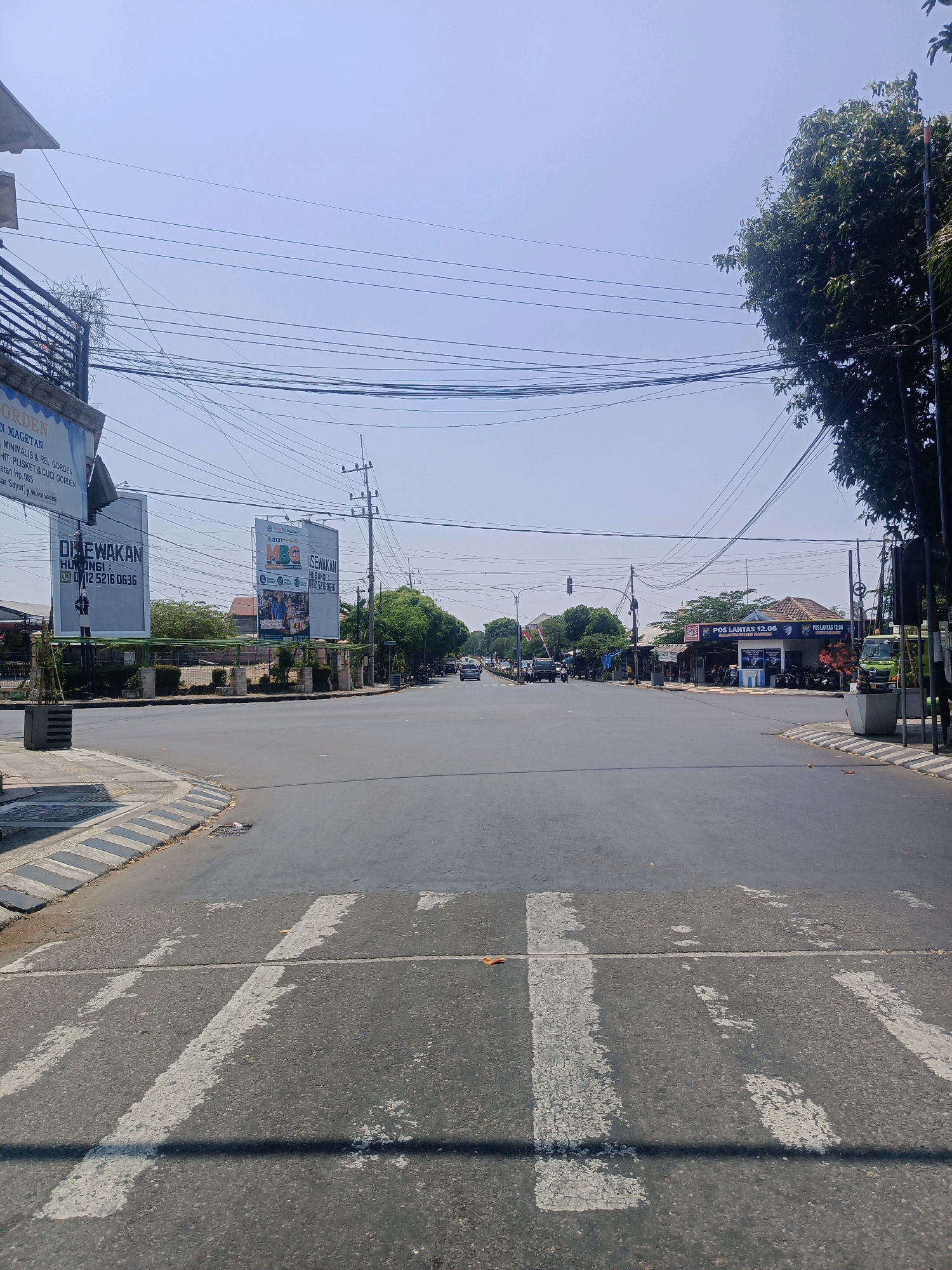 The image captures an intersection in Sukowinangun, Indonesia, under a clear, bright sky. The foreground is dominated by a road with faded white pedestrian crossing lines. To the left, a grey curb with diagonal black and white markings lines the edge of the road. Beyond the curb, a sidewalk is visible, with a potted plant.
The intersection itself is largely empty of vehicular traffic, with only a few cars and motorcycles visible in the distance, moving along the roads. Overhead, a complex network of power lines crisscrosses the sky.
On the left side of the road, a large billboard with Indonesian text is prominently displayed, along with a smaller sign. Further down the road, other buildings and signs are visible, including one that reads "POS LANTAS 12.06". To the right of the intersection, a lush green tree with dense foliage partially obscures the sky, and a dark pole stands nearby. Several parked motorcycles are visible in front of a building on the right. The overall impression is a typical street scene in a developed area of Indonesia.