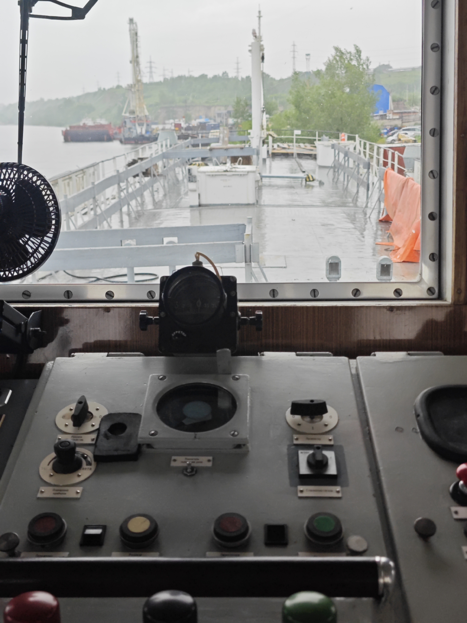 The image depicts the interior of a ship's bridge, looking out at a cloudy, overcast sky and a body of water. A complex control panel with various buttons, dials, and switches is in the foreground. In the background, a ship with a crane is docked at a port. The scene appears to be in an industrial area, possibly along the Yenisei River in Krasnoyarsk, Russia, given the presence of the river, industrial cranes, and ships commonly found in the region. The weather is damp, with water visible on the deck of the docked ship and on the window panes.