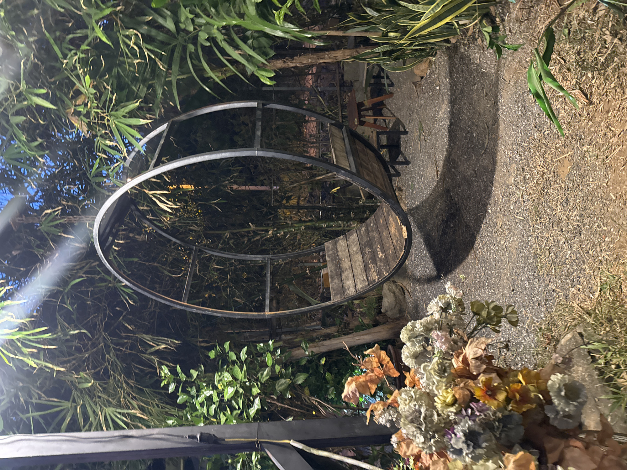 A slightly tilted long shot captures a circular metal structure with wooden planks inside, set amidst lush greenery and foliage. The structure appears to be a type of walkway or decorative feature, surrounded by various plants, including bamboo-like leaves and blooming flowers in the foreground. The ground is covered in gravel, and a glimpse of outdoor seating can be seen in the background. The image suggests an outdoor garden or park setting, possibly in Đà Nẵng, Vietnam, given the tropical flora. The scene is captured at dusk or in dim lighting, casting shadows and giving it a slightly mysterious atmosphere.