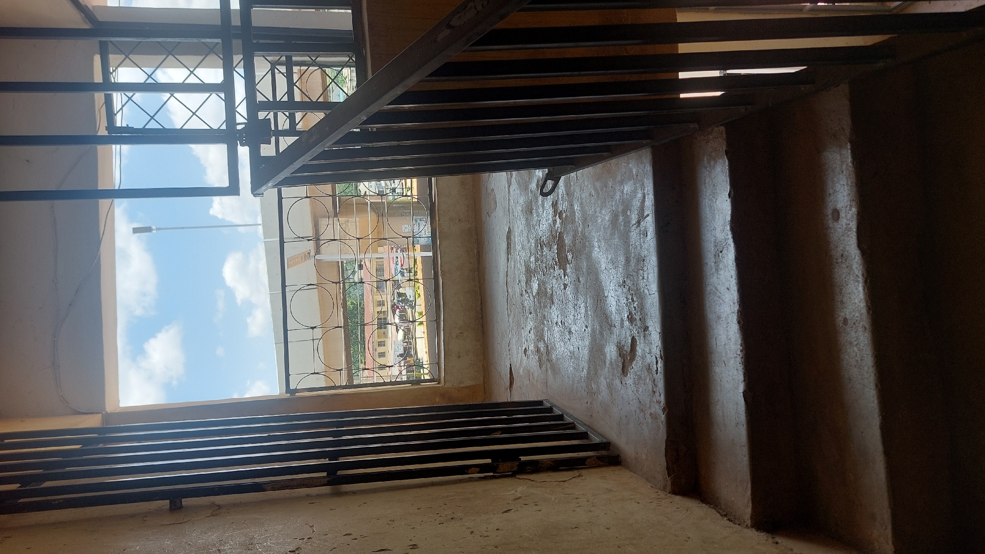 The image shows a concrete staircase leading up to a landing with a view of an outdoor environment. The landing features a metal railing and a window with a circular metal grate pattern. Through the window, buildings, vehicles, and trees are visible under a partly cloudy sky. The walls of the stairwell are a light brown color, and the concrete floor of the landing appears worn. The overall scene suggests an outdoor public space, potentially an elevated walkway or a building entrance, located in Jos, Nigeria.