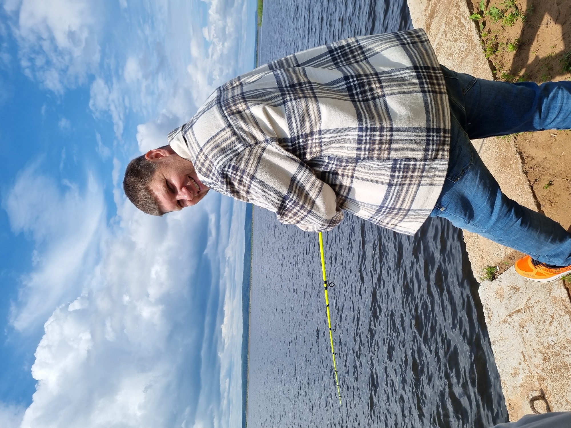 A man is pictured from the waist down, holding a fishing rod. He is wearing a plaid shirt, blue jeans, and orange sneakers. He is standing on a concrete ledge next to a body of water. The sky is blue with white clouds, and there is a distant shoreline visible across the water. The scene appears to be set in Chaikovsky, Russia, near the Kama River.