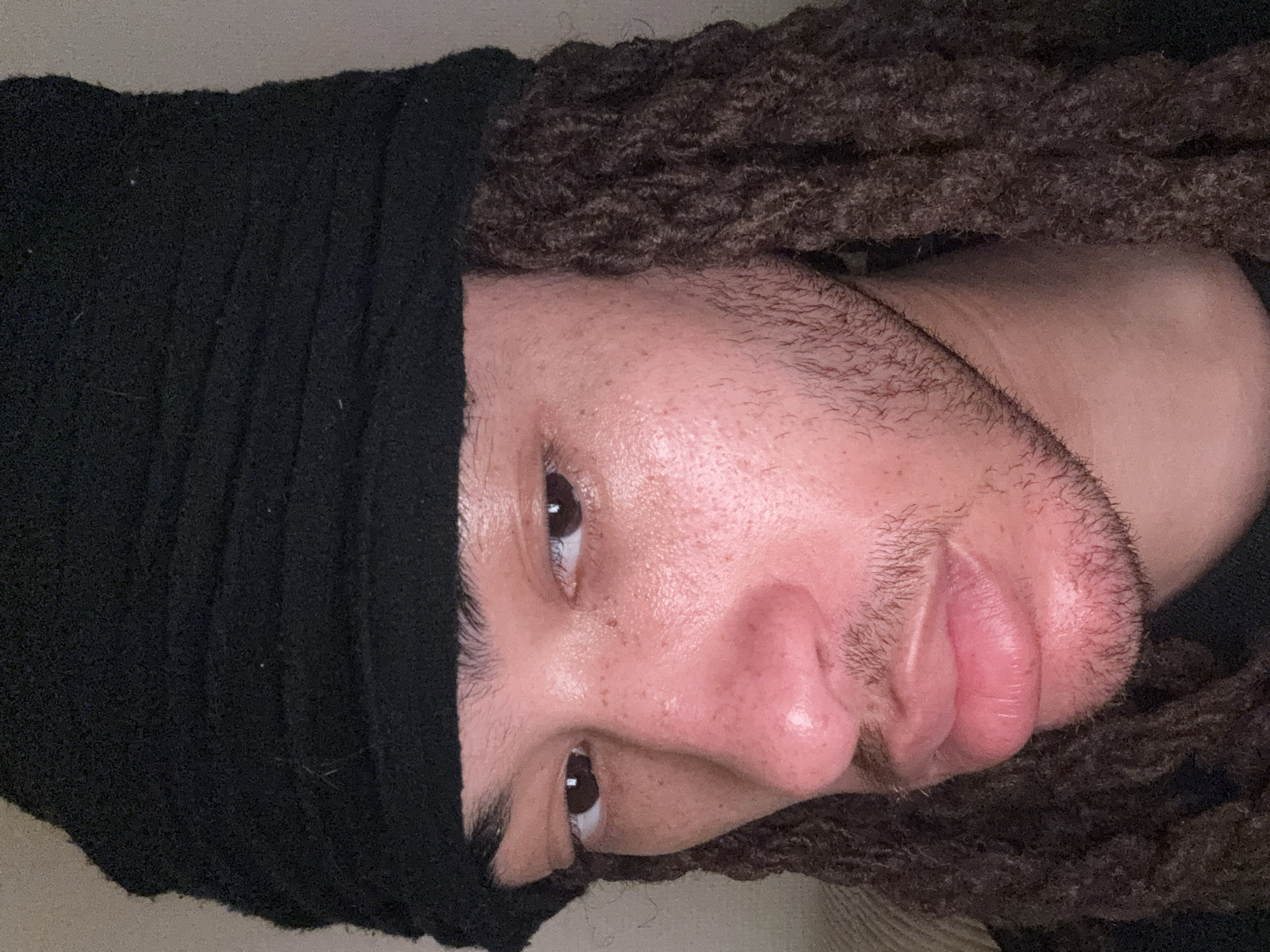 A close-up headshot of a person with dark brown eyes and a black head covering. The person has brown dreadlocks framing their face. The background appears to be a neutral tone, possibly a wall or fabric. The setting is impossible to determine and nothing indicates Paris, France.