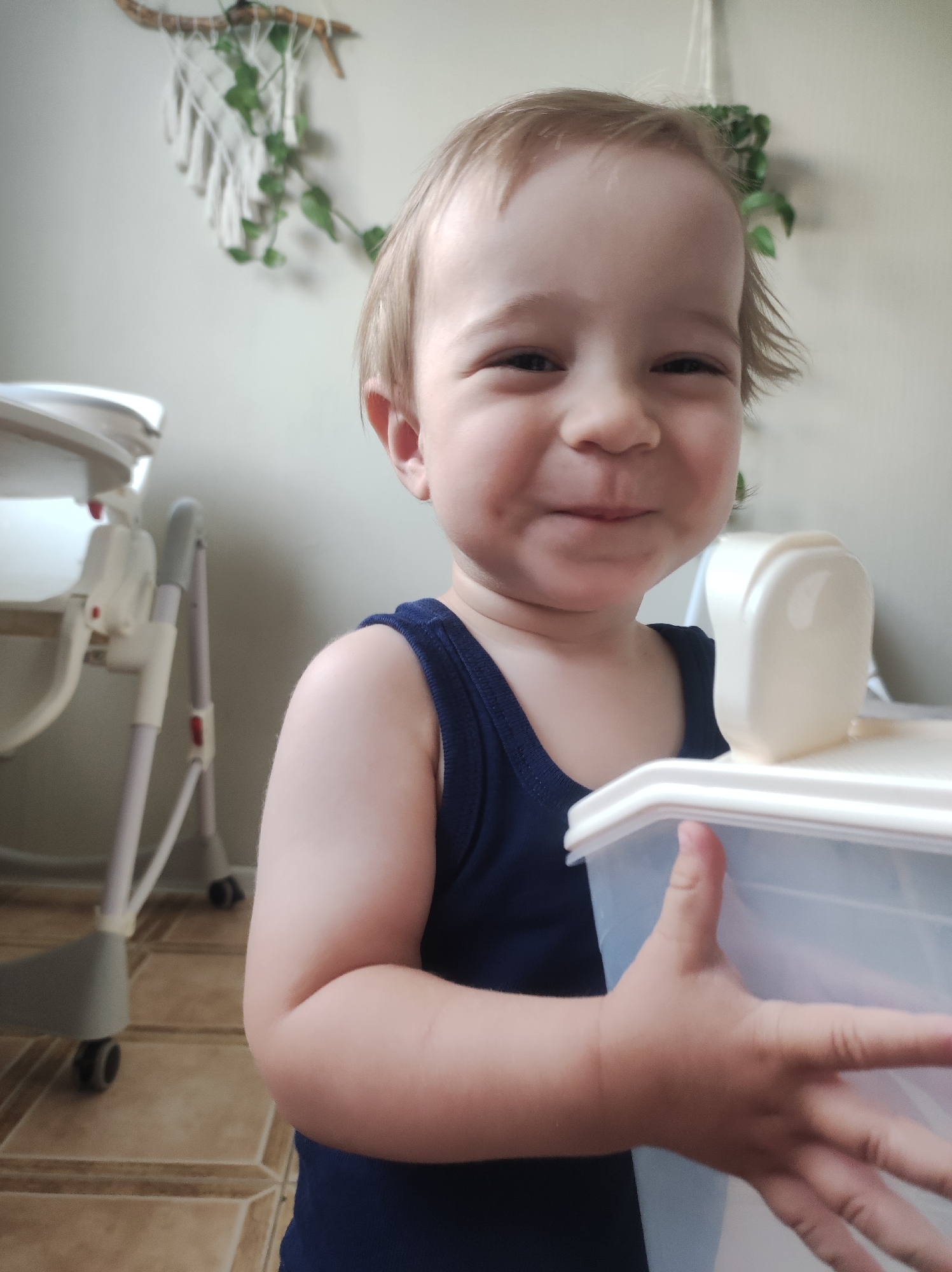 A young child with fair skin and light blond hair is smiling and holding a translucent white plastic container with a white lid. The child is wearing a dark blue sleeveless shirt. Behind the child is a high chair on the left and a wall decoration with green leaves on the right. The floor has a tiled pattern. The location appears to be indoors. There is no indication of the location being in Chaikovsky, Russia.
