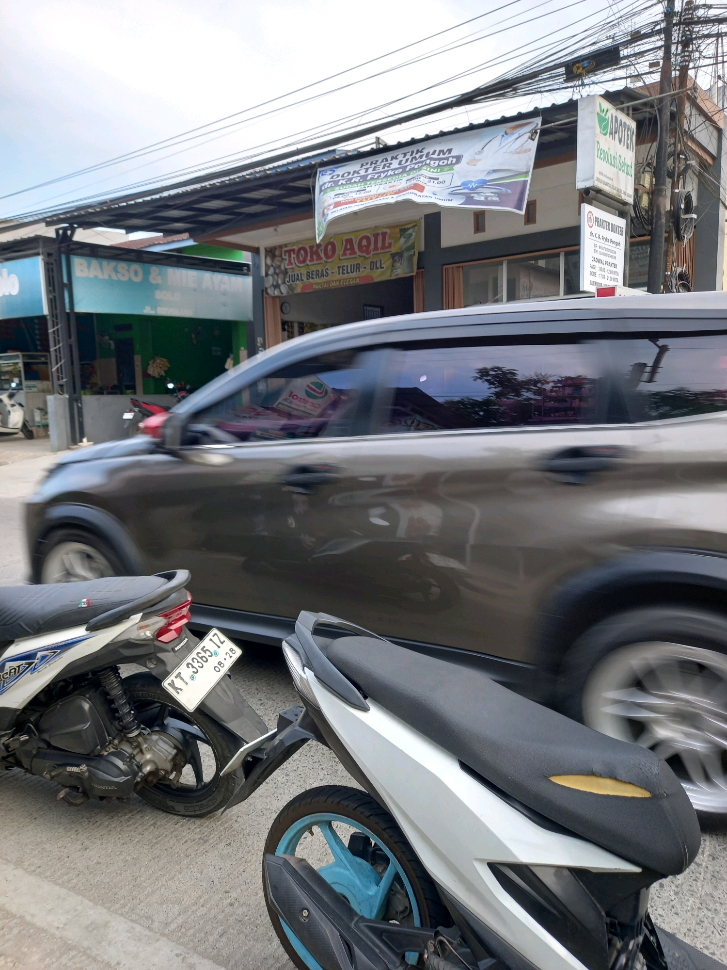The image captures a street scene in Samarinda, Indonesia, on a bright, overcast day. In the foreground, the rear sections of two motorcycles are visible. The left motorcycle is white with blue accents, and its license plate reads "KT 3365 1Z 08-28". The right motorcycle is partially obscured but shows a blue wheel rim and black accents.
A dark-colored car is moving in the middle ground, its motion blurred by the camera's shutter speed. The car's tinted windows reflect the sky and surrounding buildings.
In the background, a row of commercial establishments lines the street. Signs indicate a "BAKSO & MIE AYAM" (meatball and noodle soup) shop and a "TOKO AQIL" which sells rice and other items. Further down the street, an "APOTEK Revolusi Sehat" (Healthy Revolution Pharmacy) and a "PRAKTEN DOKTER" (Doctor's Practice) are advertised with signage including doctor names and operating hours. Electrical wires are strung overhead, typical of an urban environment in Indonesia. The overall impression is a bustling street scene in a commercial area of Samarinda.