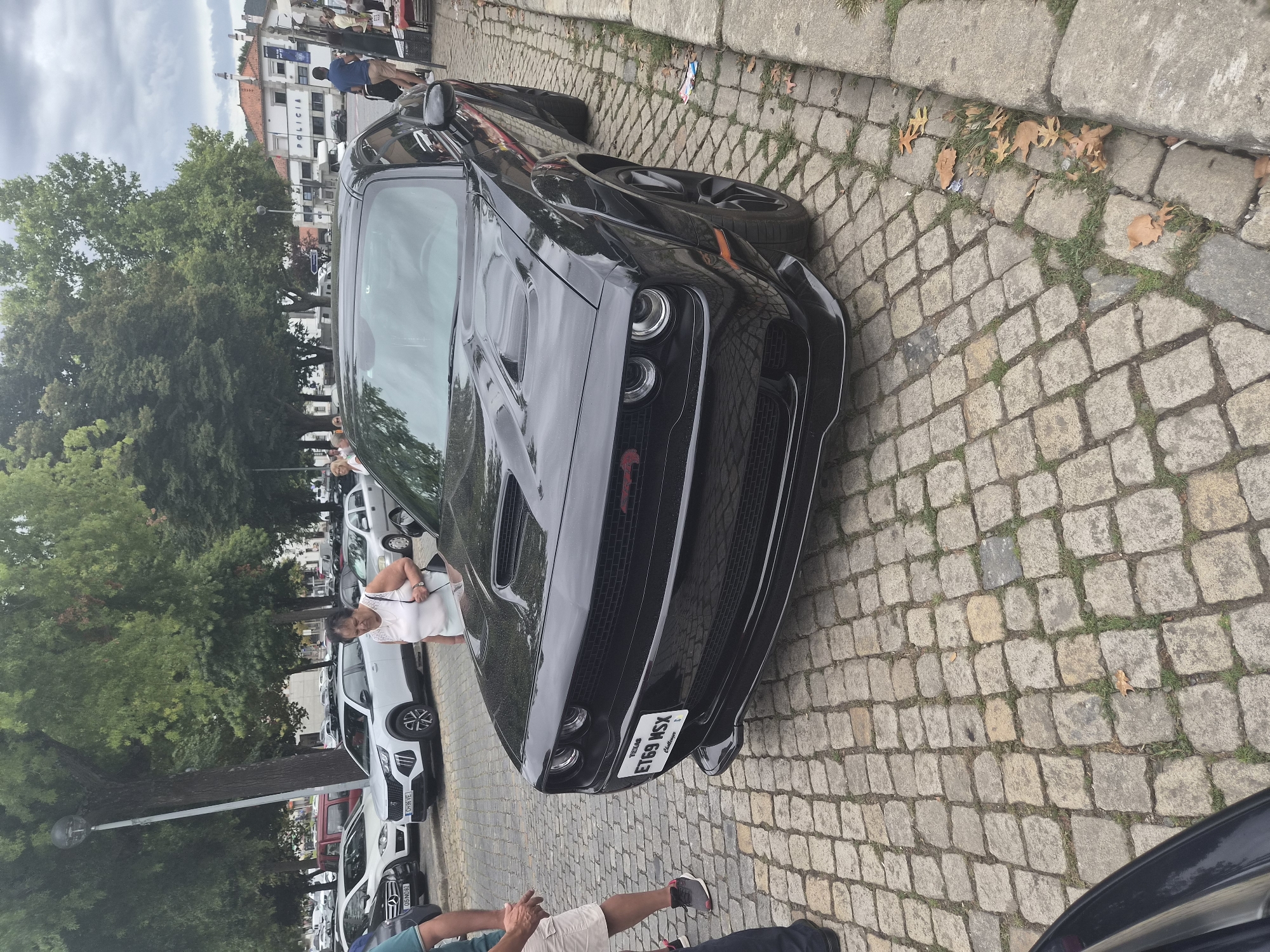 A black Dodge Challenger is parked on a cobblestone street in Barcelos, Portugal, in front of a building labeled "POLICIA." The car's license plate reads "ET69 MSX." A woman in a white top and dark pants is walking past the car. Several other cars are parked in the background, and trees line the street. The sky is overcast.