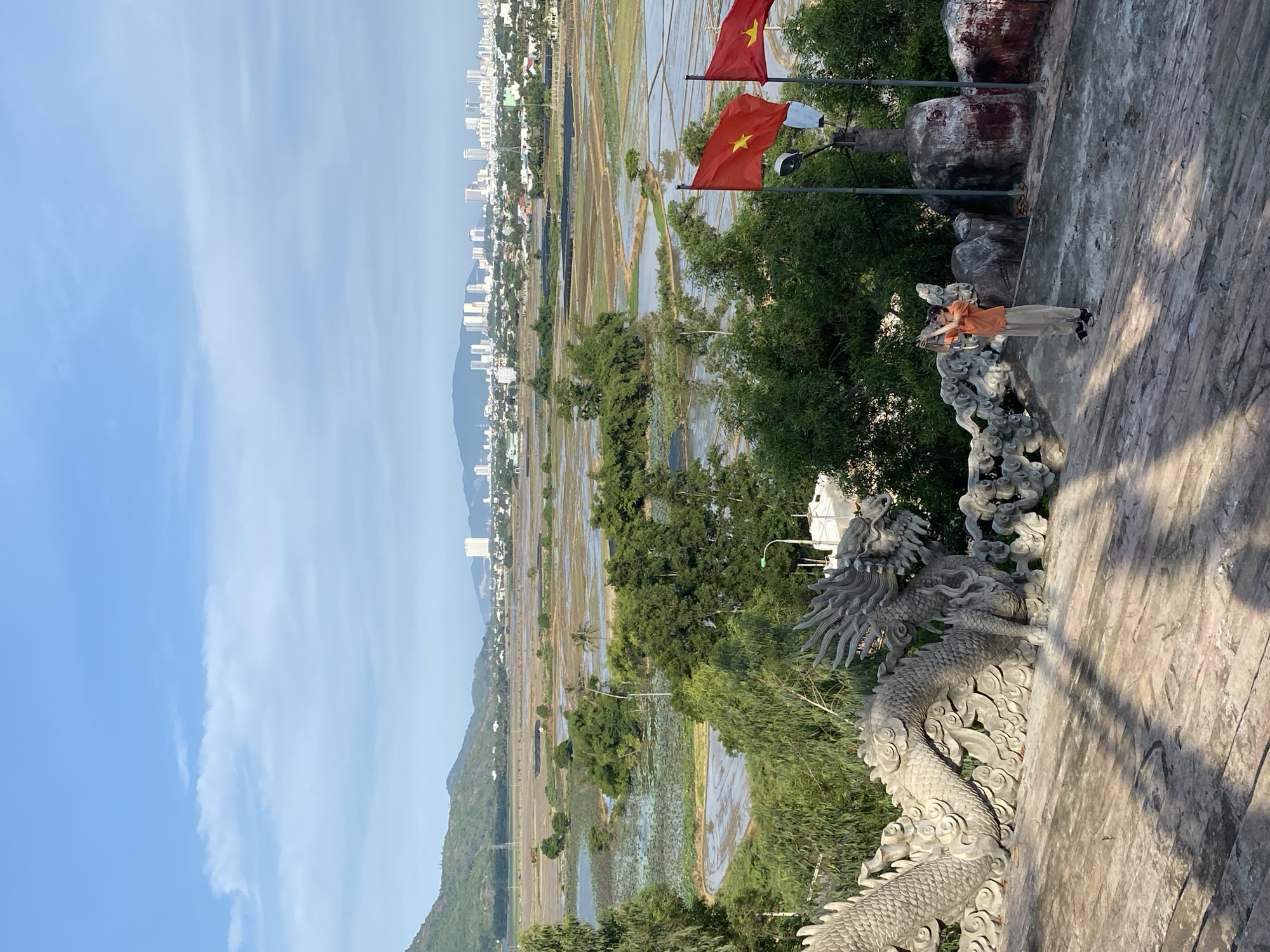 A full shot shows a person standing on a stone stairway, holding a camera up to their face as if taking a photograph. The person is wearing a light orange shirt and tan pants. In the foreground, a stone dragon statue with intricate details lines the edge of the stairway. Behind the person, two Vietnamese flags wave on poles. In the background, a panoramic view of the city of Tay Nha Trang, Vietnam, unfolds with numerous buildings, some of which are tall high-rises. The city is nestled between a large body of water, possibly a bay or estuary, and lush green hills. The sky is clear with a few wispy clouds. The overall scene suggests a scenic viewpoint overlooking a coastal city.