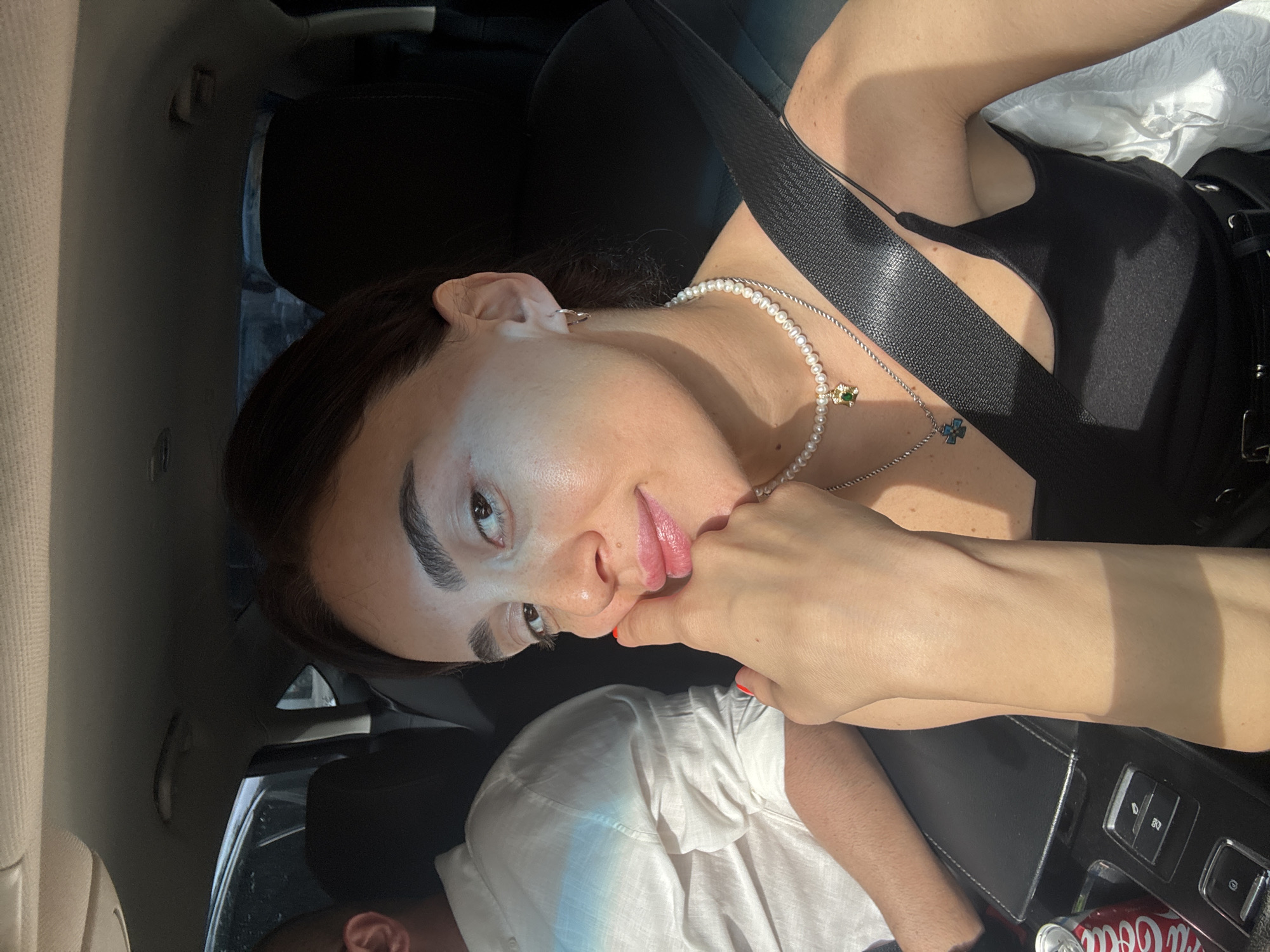 A woman with dark hair, wearing a black tank top and a pearl necklace, sits in the passenger seat of a car. She is looking at the camera and resting her chin on her hand. The woman has on hoop earrings and her nails are painted red. A man wearing a white collared shirt is visible in the background, to her left. A can of Coca-Cola sits in the car's center console. The interior of the car appears to be dark, with sunlight streaming in from the side. The scene suggests a casual setting, possibly a road trip. The image does not provide enough information to determine the specific location within the city of Tbilisi, Georgia.