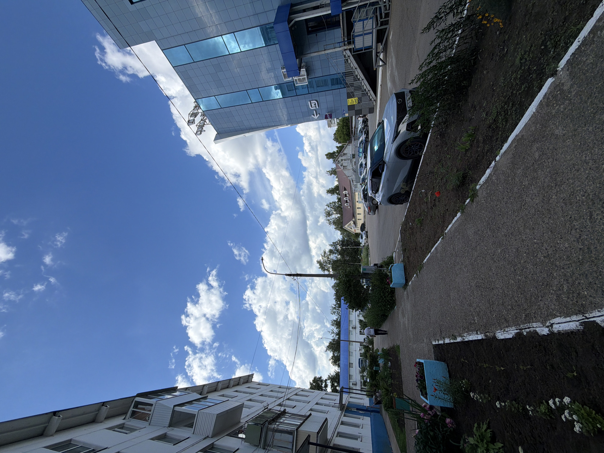 The image depicts a street scene in Chaikovsky, Russia, on a bright, cloudy day. On the left, a multi-story apartment building with white and blue accents dominates the foreground. A paved walkway runs alongside the building, bordered by garden beds with plants and blue planters. A lone figure in a white shirt and dark pants walks away from the camera down this path.
To the right of the walkway is a paved area with parked cars. A silver SUV is prominently positioned in the foreground. Further back, other vehicles are visible, including a red sedan and a dark-colored SUV. In the mid-ground, a streetlamp stands tall, with overhead wires stretching across the scene. Beyond the parked cars, a building with a distinctive blue, modern facade and large glass windows stands out. This building appears to be a commercial or public facility, with stairs leading up to an entrance. Adjacent to this structure, a low-rise building with a blue roof is visible. In the background, a cluster of houses with peaked roofs and trees complete the urban landscape under a blue sky with scattered white clouds.