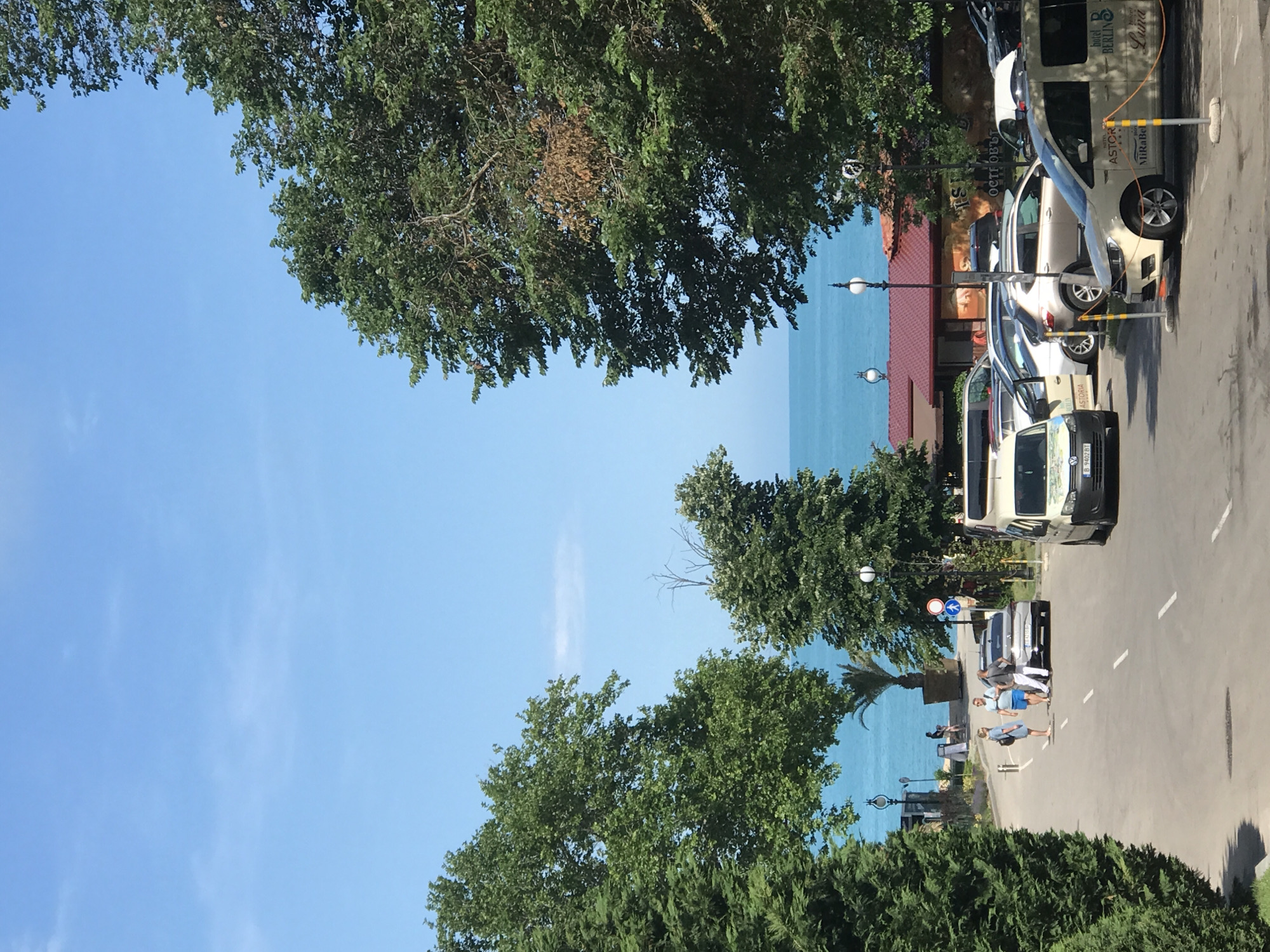 The image depicts a sunny day in the city of Varna, Bulgaria, specifically along the Sea Garden, overlooking the Black Sea. The scene is a paved road with several parked vehicles, including a white van with "HOTEL ASTOR" and "BERLIN MiraBella" written on its side, a silver sedan, and other cars. Three people are standing on the road, engaged in conversation. One person is wearing a blue dress, another a light blue t-shirt and shorts, and the third a grey t-shirt and dark pants. Further down the road, more people are visible near the coastline, and there are buildings with red roofs and outdoor seating areas. Lush green trees frame the scene, with the vibrant blue sea and sky dominating the background. The overall impression is a peaceful, summery atmosphere in a coastal city.