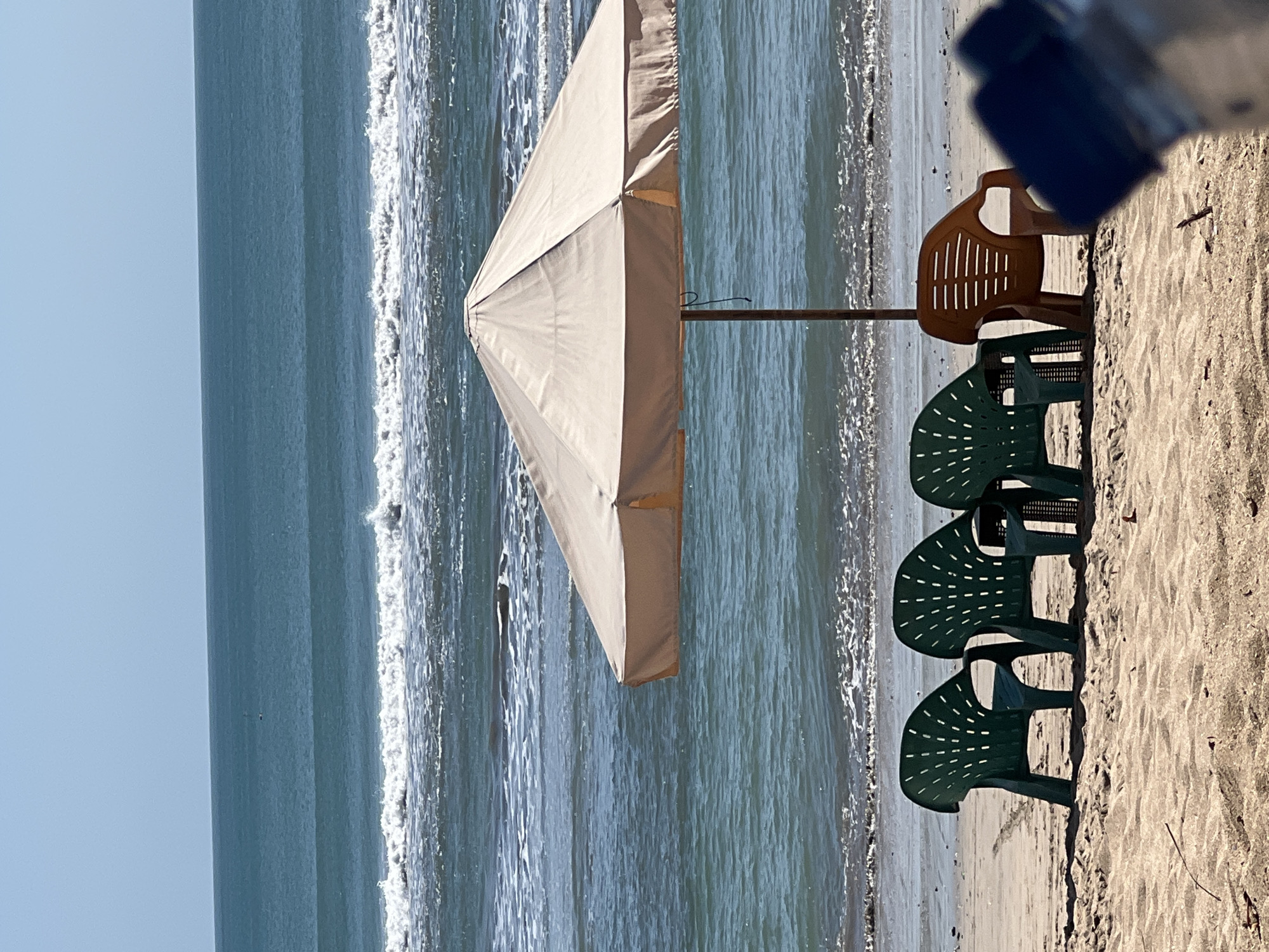 The image depicts a beach scene in Kuta, Indonesia, with the ocean in the background. In the foreground, the sand is visible with several beach chairs and an umbrella. Three dark green plastic chairs are arranged in a row, facing the ocean. To the right of these, a single orange plastic chair is positioned. A beige umbrella with a metal pole stands behind the chairs, providing shade. The ocean water is a deep blue-green, with gentle waves breaking on the shore. The sky above is clear and blue. In the far distance, on the water, a few faint dark shapes are visible, possibly seabirds or small boats. The overall impression is a serene and inviting beach setting.