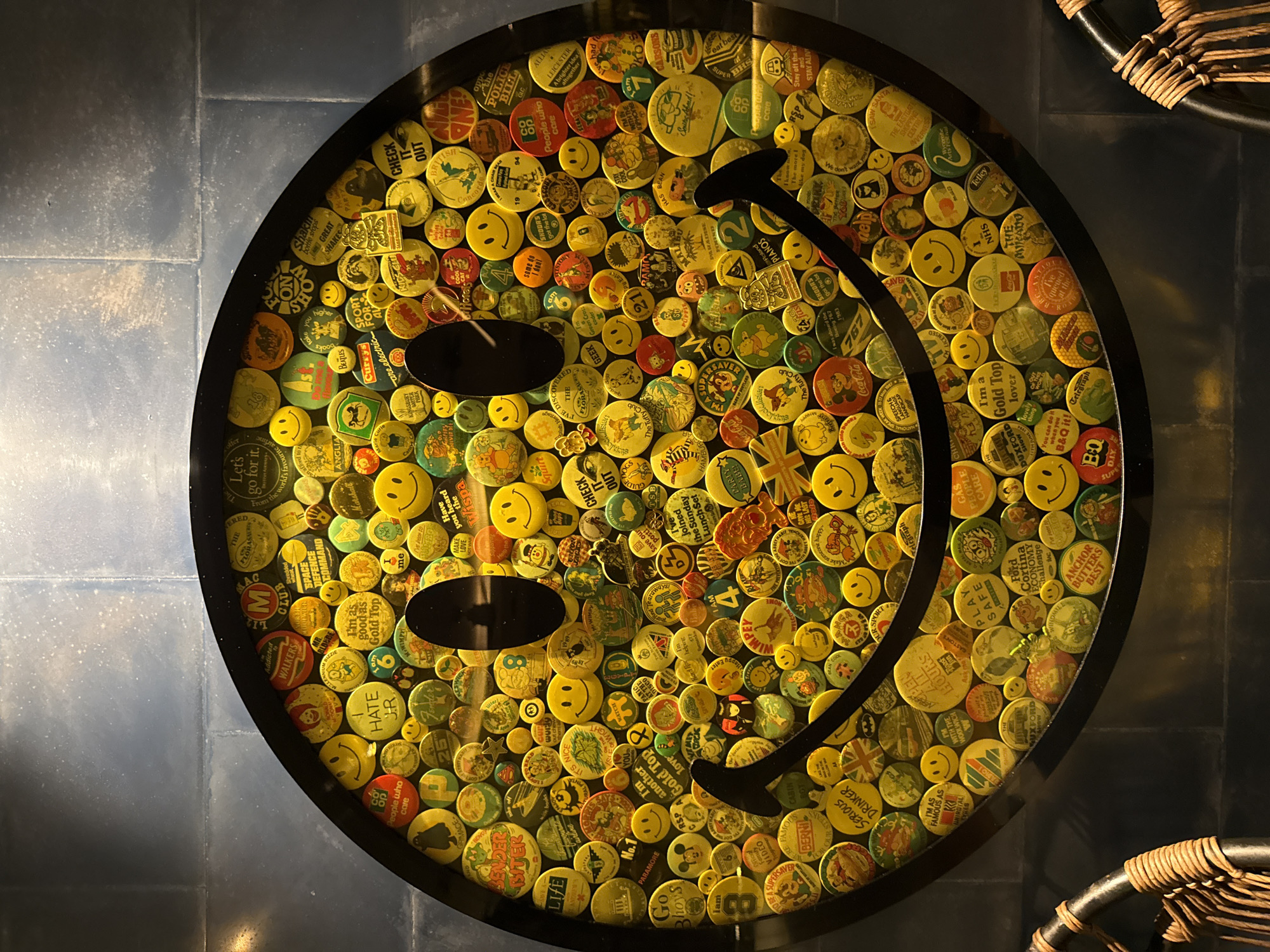 A circular black table in Kuta, Indonesia, is visible from an overhead perspective. The table’s surface is decorated with a dense collection of yellow and other colored circular buttons, arranged to form a stylized smiley face. Two horizontally-oriented black ovals represent the eyes, while a curved black line creates the smile. The background features dark gray, square floor tiles, with wicker furniture partially visible at the edges of the frame.