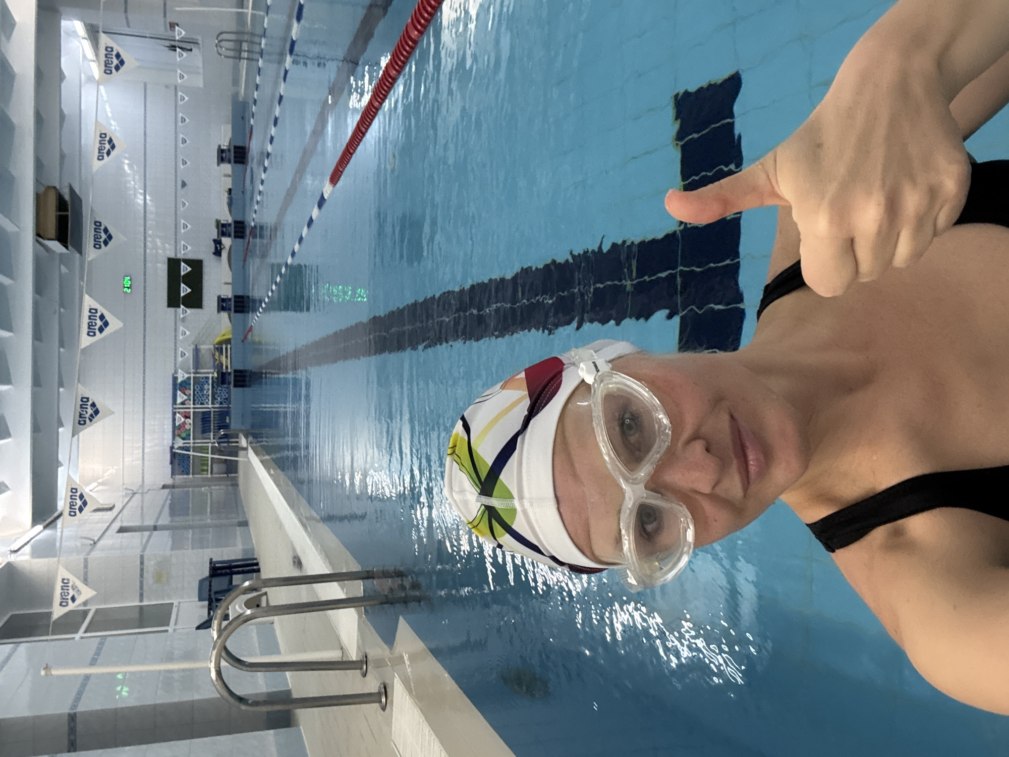 The image shows a woman in a swimming pool. She is wearing a swim cap and goggles, and giving a thumbs-up. The pool is indoors and has lane markers. Pennants with the "Arena" logo are visible along the side of the pool. Based on the location, this is likely a swimming pool located in the city of Chaikovsky, Russia.