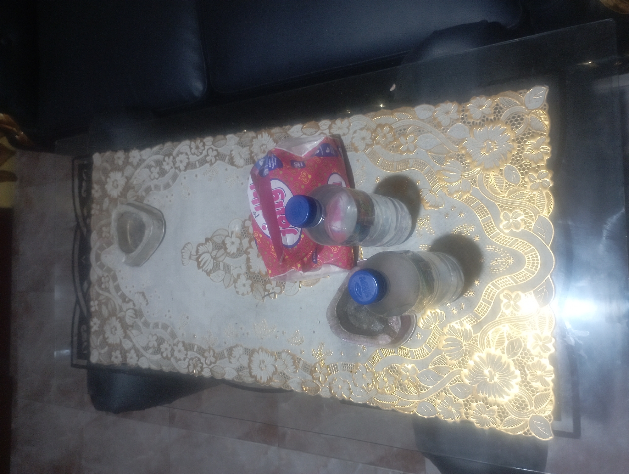 An overhead shot shows a coffee table in Maos Kidul, Indonesia, covered by a decorative lace tablecloth with floral patterns and gold trim. On the table are two plastic water bottles, one with a blue cap and the other partially obscured, and a package of pink wet wipes with the brand name "Jolly" visible. A small, gray ashtray or dish is also present. The table appears to be made of glass and is positioned next to a dark leather sofa, part of which is visible in the frame. The surrounding environment is dimly lit, with some light reflecting off the table surface.