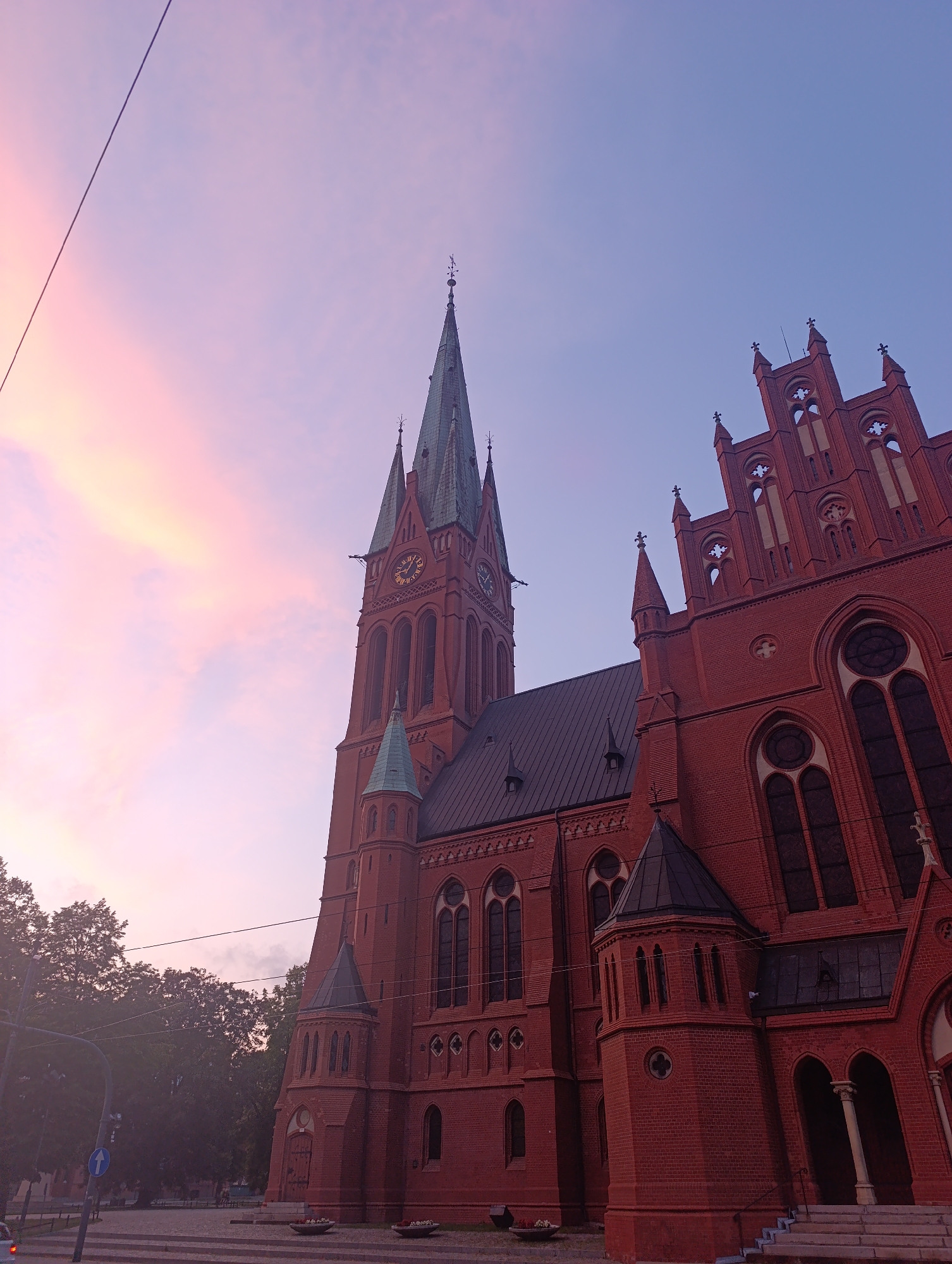The image shows the Holy Ghost Church, a historic red-brick Gothic Revival church located in Toruń, Poland, during a twilight sky. The church features tall spires, arched windows, and intricate architectural details. The sky above is a gradient of soft pink and purple hues, indicating either dawn or dusk. A wire runs diagonally across the upper left portion of the frame. In the lower left, trees and street elements, including a traffic signal and a street sign, are visible, suggesting an urban setting. There are no people or specific actions depicted in the scene.