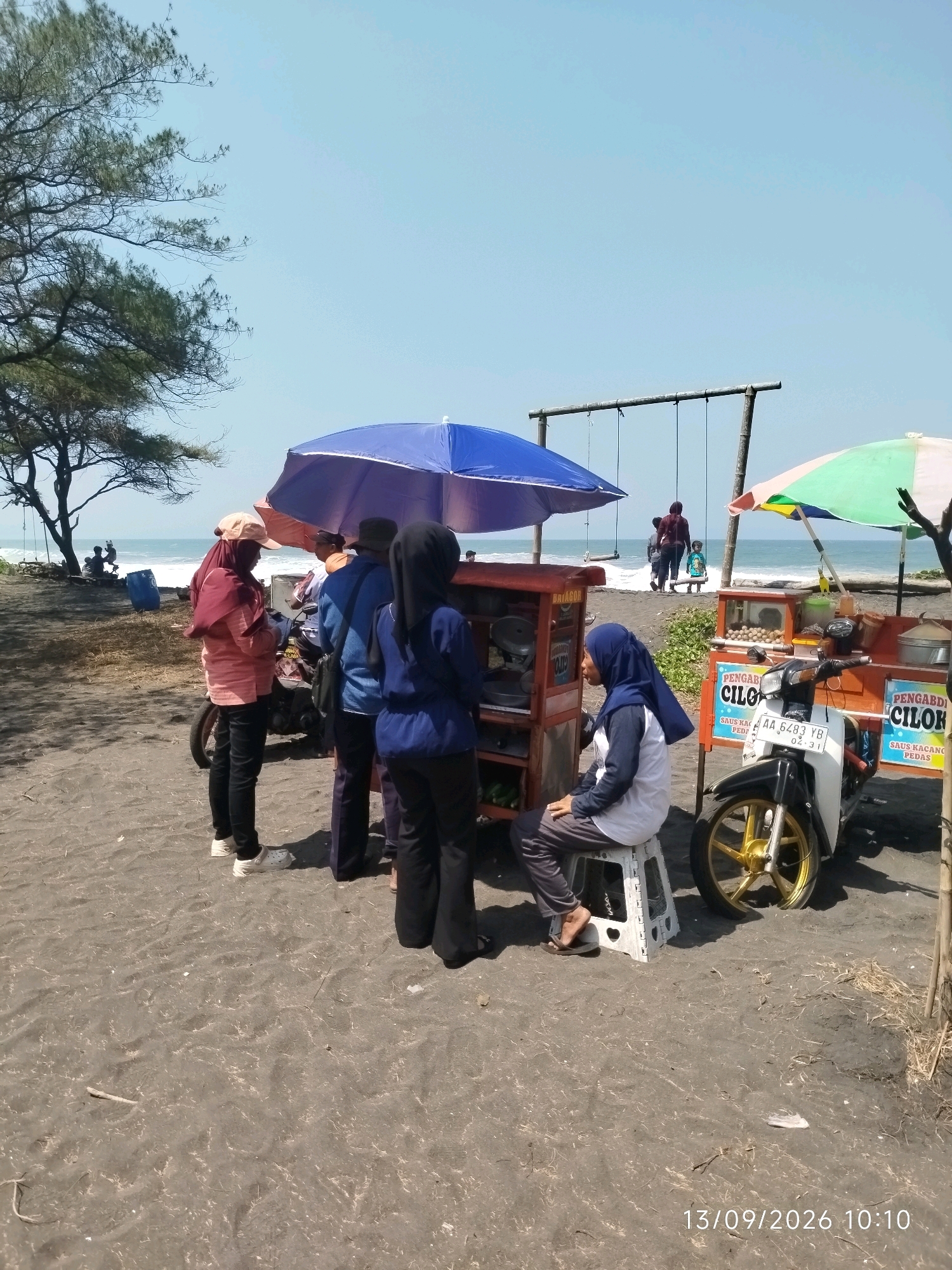 The image depicts a beach scene in Pleret, Indonesia, on a sunny day with a clear blue sky. In the foreground, a black sand beach is visible, with some dried vegetation scattered across it. Several people are present, including a woman wearing a pink headscarf and a peach-colored shirt, standing near a food cart. Another person, also wearing a headscarf, is sitting on a white foldable stool next to the cart. A third individual, in a dark blue headscarf and shirt, stands near the cart. A motorcycle with a golden rim is parked in front of one of the carts. Two food carts are visible: one red and one orange. The red cart has a sign that reads "BATAGOR". The orange cart has signs that say "PENGAD" and "CILOK", with smaller text below indicating "SAUS KACANG PEDAS". A large blue umbrella and a smaller multi-colored umbrella provide shade over the carts. In the background, a swing set is visible, and further back, people are walking along the shoreline where waves are breaking. A large tree with sparse foliage stands to the left of the scene. A blue plastic barrel is located near the tree. The date and time stamp "13/09/2026 10:10" is present in the bottom right corner of the image.