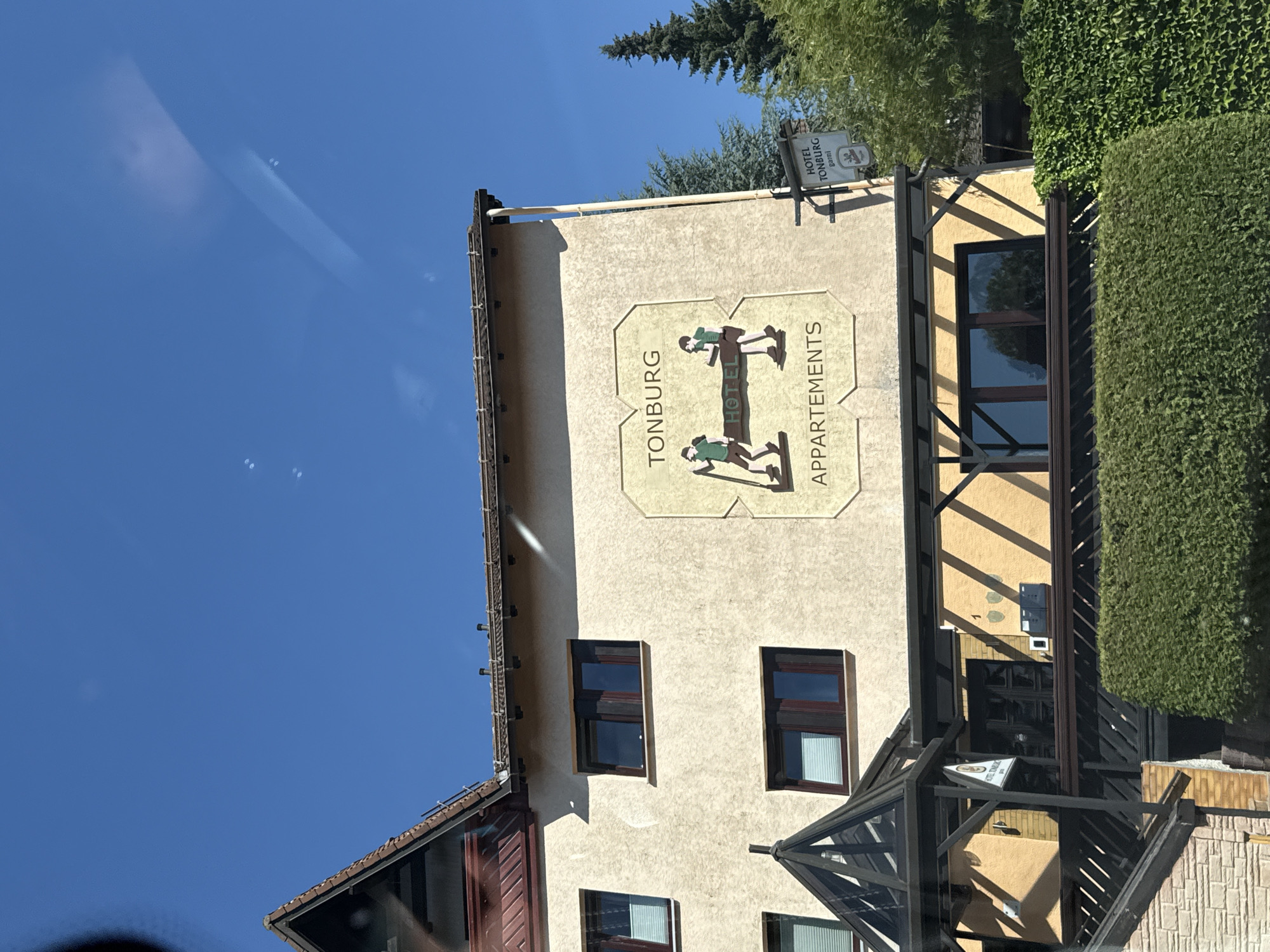 The image shows the Hotel Tonburg in Bad Sachsa, Germany. The hotel is a light-colored building with several windows. A large sign on the side of the building reads "TONBURG APPARTEMENTS" with an illustration of two figures carrying a long pole. There is also a smaller sign that says "HOTEL TONBURG". The hotel is surrounded by trees and greenery. The sky is blue and clear.