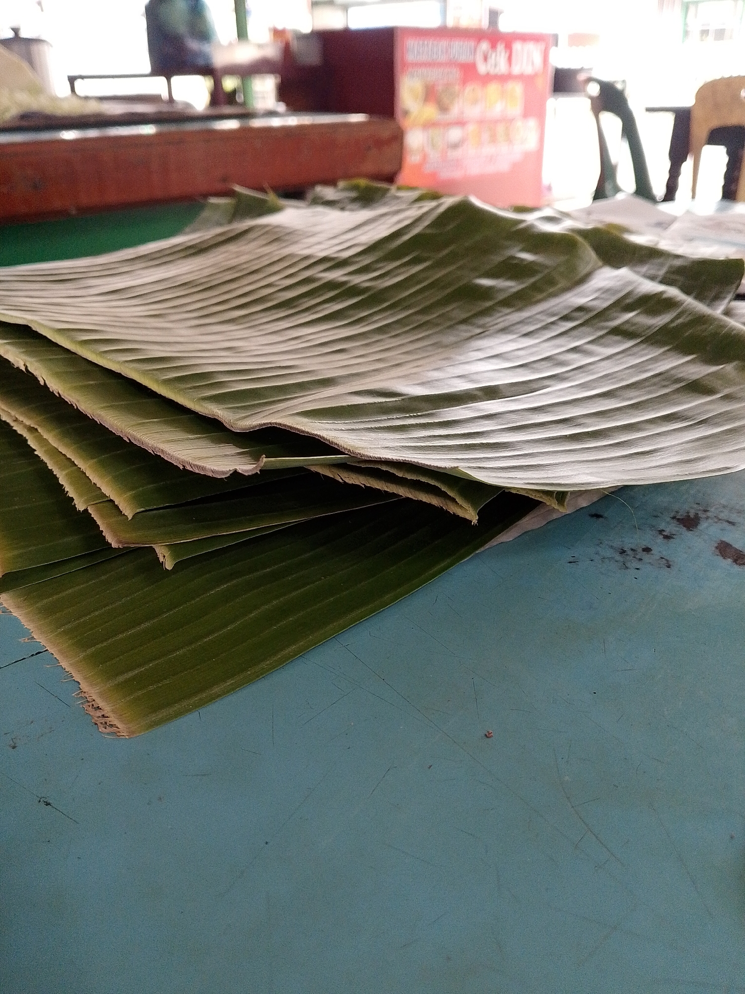 A stack of large, green banana leaves lies on a turquoise-colored table in the city of Lhokseumawe, Indonesia. The leaves are curved and ribbed, with their edges appearing slightly dried and frayed. In the background, a reddish-brown counter is visible, behind which a blurred figure is seated. Further in the background, a brightly colored sign with text in a foreign language is visible, along with other blurred objects and furniture. The lighting suggests an indoor setting, possibly a food stall or market.