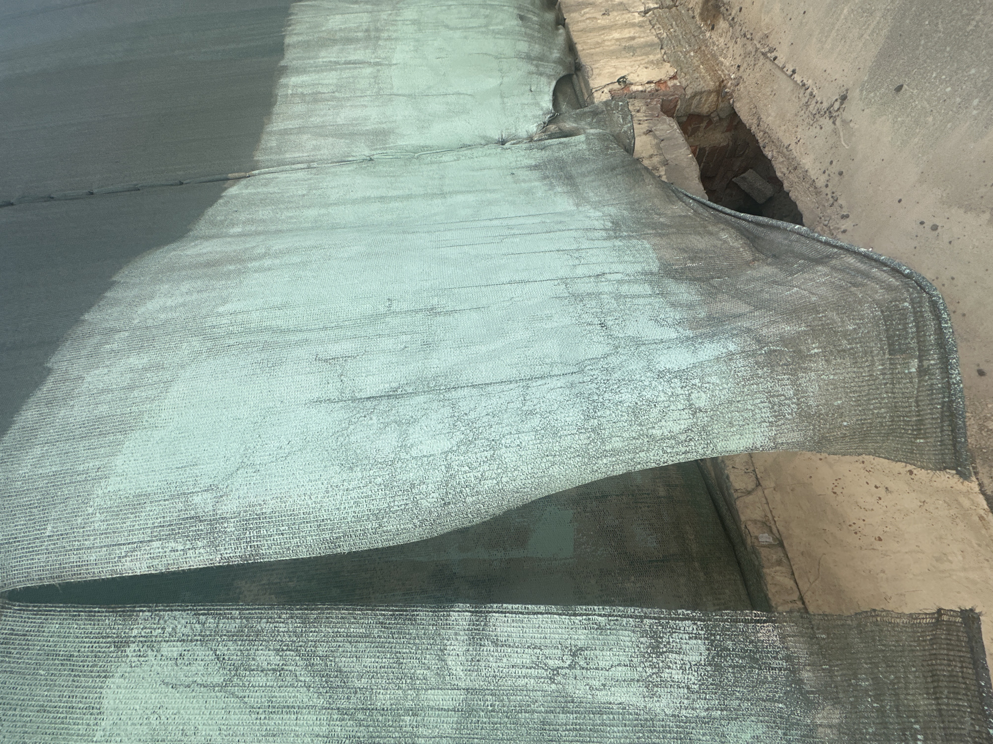 The image shows a close-up view of a green mesh or netting partially covering a concrete structure. The netting appears to be layered or draped, with some sections raised or overlapping others. To the right of the frame is a section of concrete wall with signs of deterioration, including exposed brick. A dark recess or hole is visible within the concrete structure. The setting is consistent with a construction site in Saint Petersburg, Russia.
