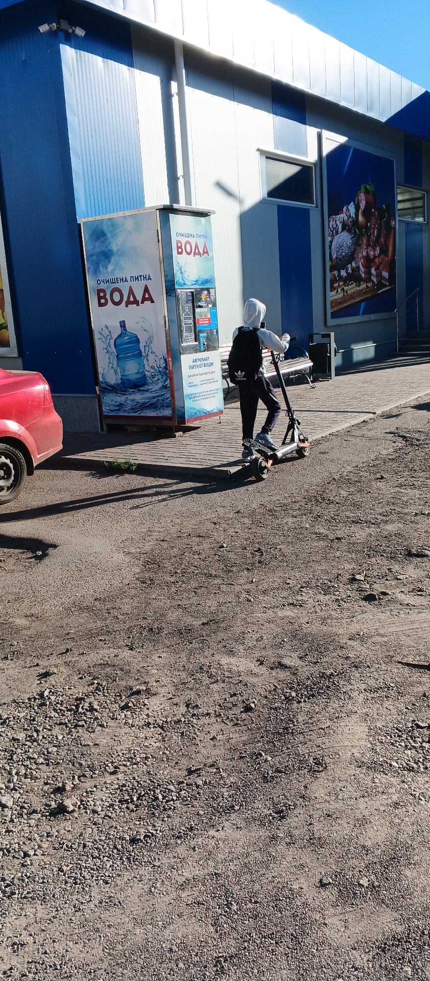 In the city of Smila, Ukraine, on a sunny day, a young person wearing a light-colored hooded sweatshirt, dark pants, and a backpack with the Adidas logo rides an electric scooter on a paved walkway. To the left of the walkway is a gravel surface with tire tracks, and a portion of a red car is visible on the far left. Behind the person on the scooter is a water vending machine with a large blue bottle graphic and text that reads "ОЧИЩЕНА ПИТНА ВОДА" (Purified Drinking Water) and "АВТОМАТ ПИТНОЇ ВОДИ" (Drinking Water Vending Machine). The building behind the vending machine is a store with blue and white striped siding. A large advertisement featuring processed meats is visible on the right side of the building. The sky is clear and blue.
