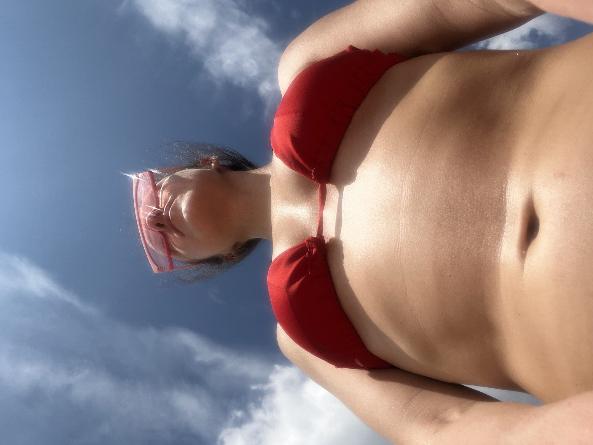 This is a low-angle, full-body shot of a person wearing a red bikini and pink sunglasses, taken outdoors against a bright blue sky with scattered clouds. The individual appears to be standing or lying down, with the camera positioned below them, looking upwards. The image is focused on the person's torso and chest, with their face partially visible at the top, looking upwards. The lighting suggests it is a sunny day. There is no visible indication of the location being in Kazan, Russia.
