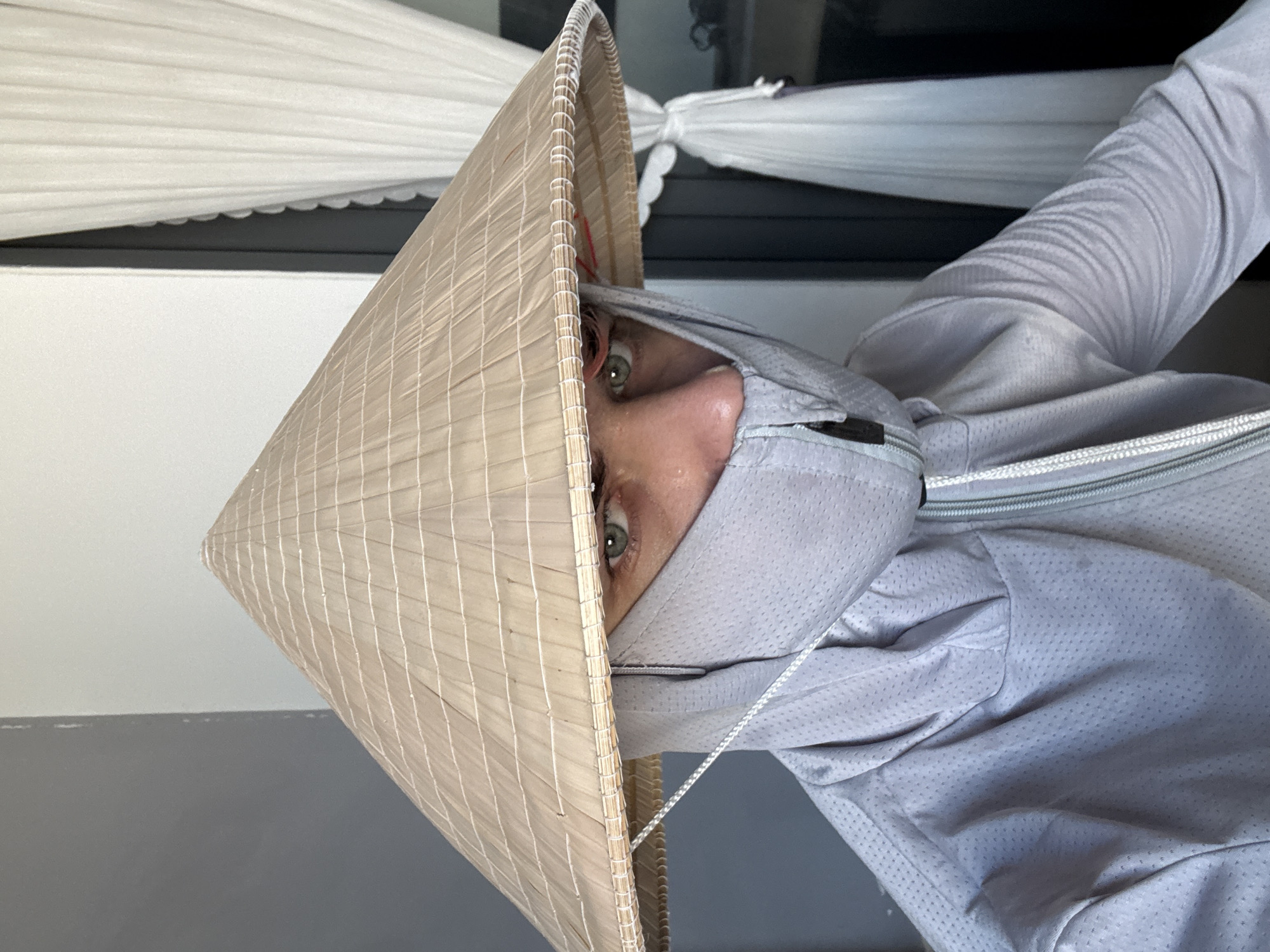 A person wearing a light gray sun protective jacket and a traditional Vietnamese conical hat is looking at the camera. The person's face is partially covered by a light gray face mask, revealing only the eyes. The hat is light brown with a woven pattern. Behind the person is a white pleated curtain and a dark window frame. The background suggests an indoor setting with natural light. The image does not provide enough information to determine the location in Đà Nẵng, Vietnam.