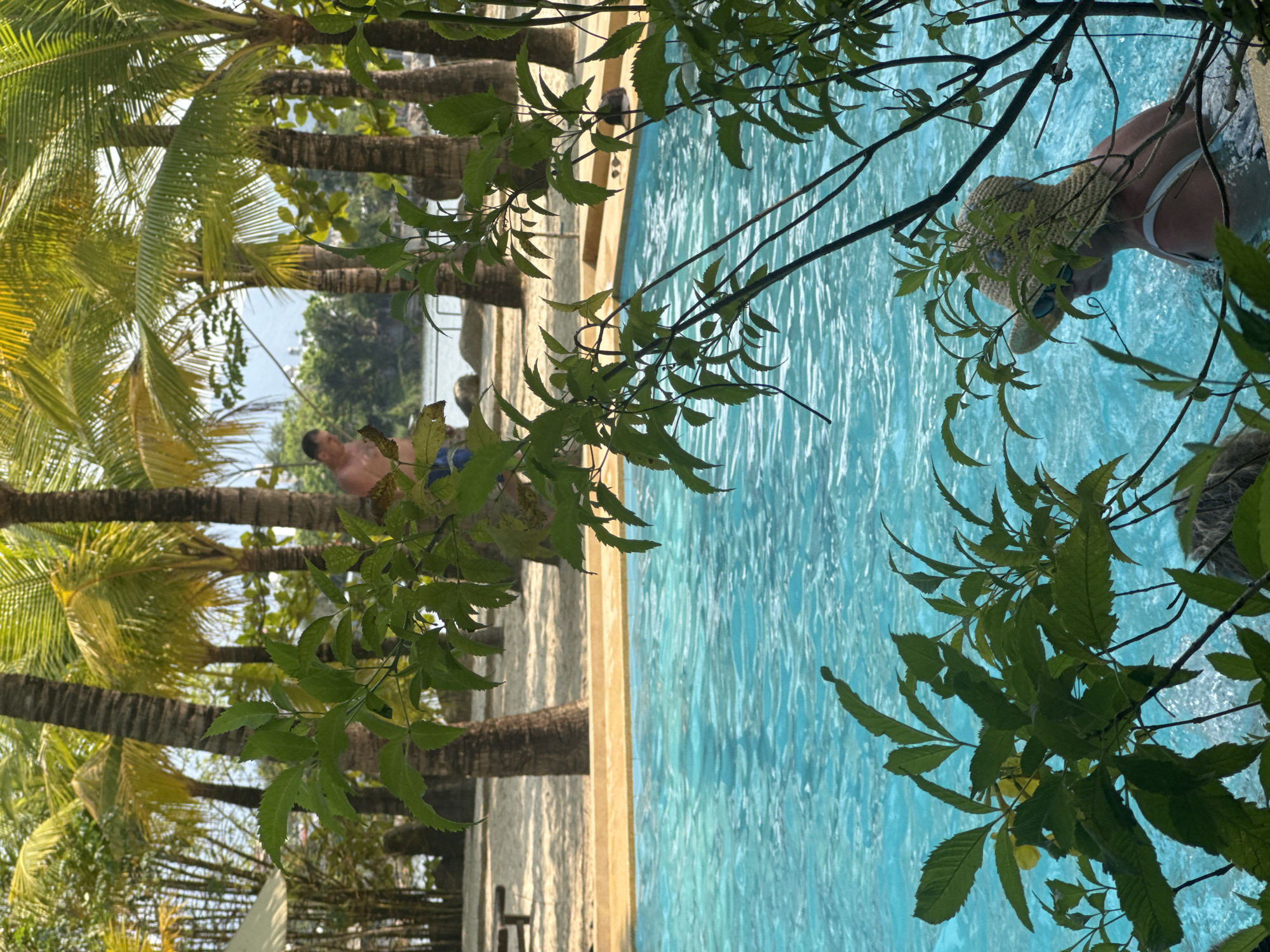 The image shows a person wearing a sunhat and sunglasses in a swimming pool. The pool is surrounded by palm trees and other greenery. A man is seen standing near the pool. The location is unknown.