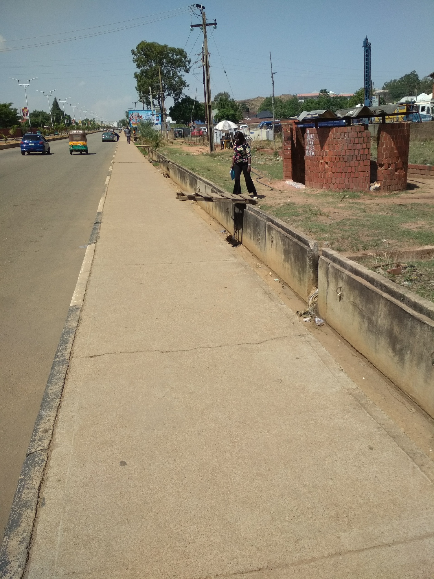 The image depicts a street scene in Barakin Lerawi, Nigeria. The view is a wide shot of a straight road with a sidewalk on the right side. On the road, a blue car and a green auto rickshaw are visible, along with other distant vehicles and pedestrians. On the sidewalk, a woman in a patterned shirt and dark pants is walking.
A concrete barrier with a drainage ditch runs along the edge of the sidewalk. Behind the barrier is a small brick structure with three openings. The area behind the structure appears to be grassy with sparse vegetation. In the background, utility poles and a drilling rig can be seen. The sky is clear and blue.