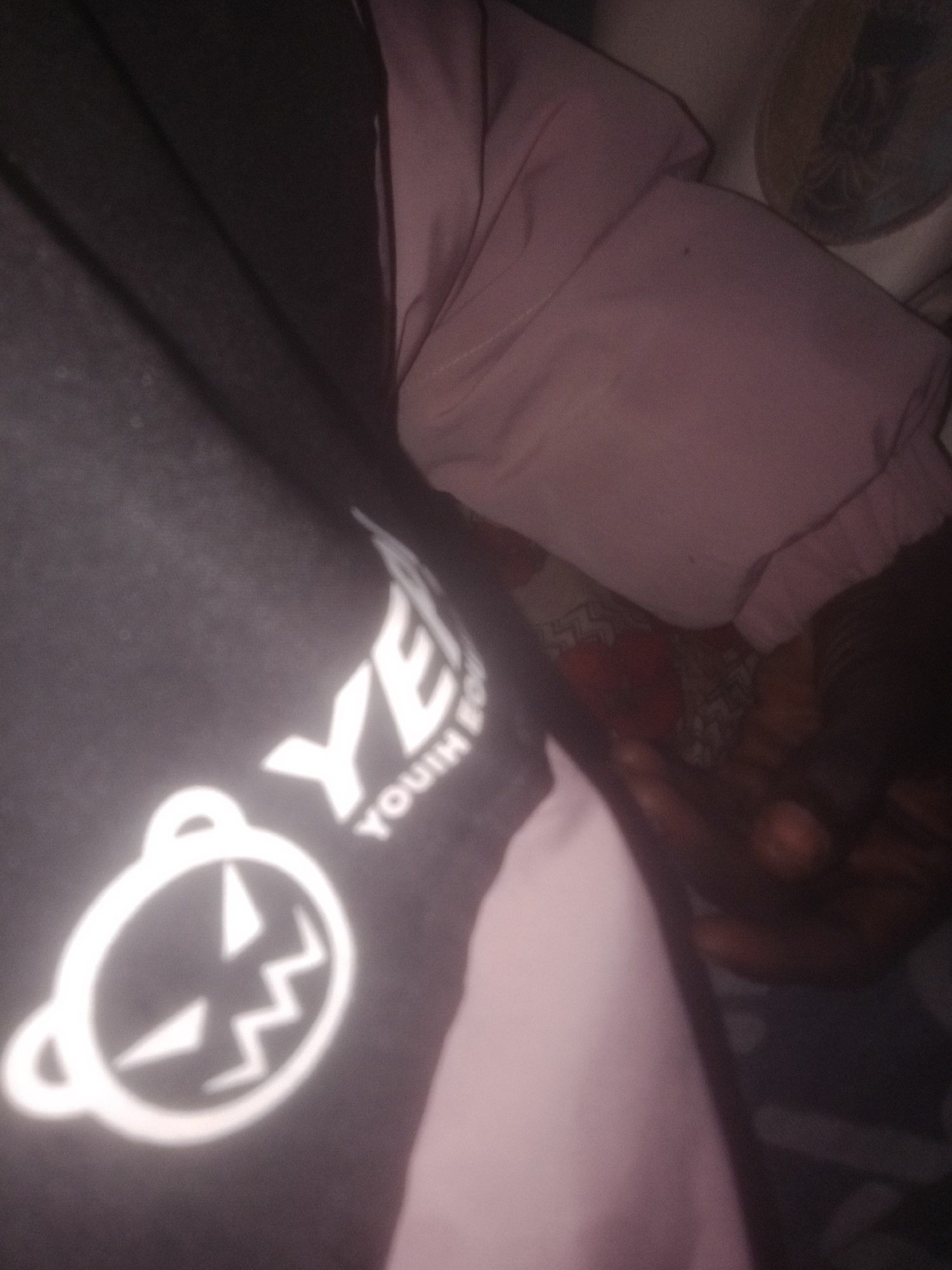 The image shows a close-up view of clothing. On the left, a black garment with a white graphic design and text is visible. The graphic is a circular outline with two ear-like protrusions at the top and a stylized, angular face with a jagged mouth inside. The text "YEH YOUIM" is partially visible next to the graphic, also in white. To the right of the black garment, a pink fabric is folded. The background is dark and out of focus, with some patterned fabric or material visible underneath the pink fold. The overall impression is of clothing laid out or draped, with a focus on the design elements. The lighting is dim, creating shadows and highlighting the white graphics.