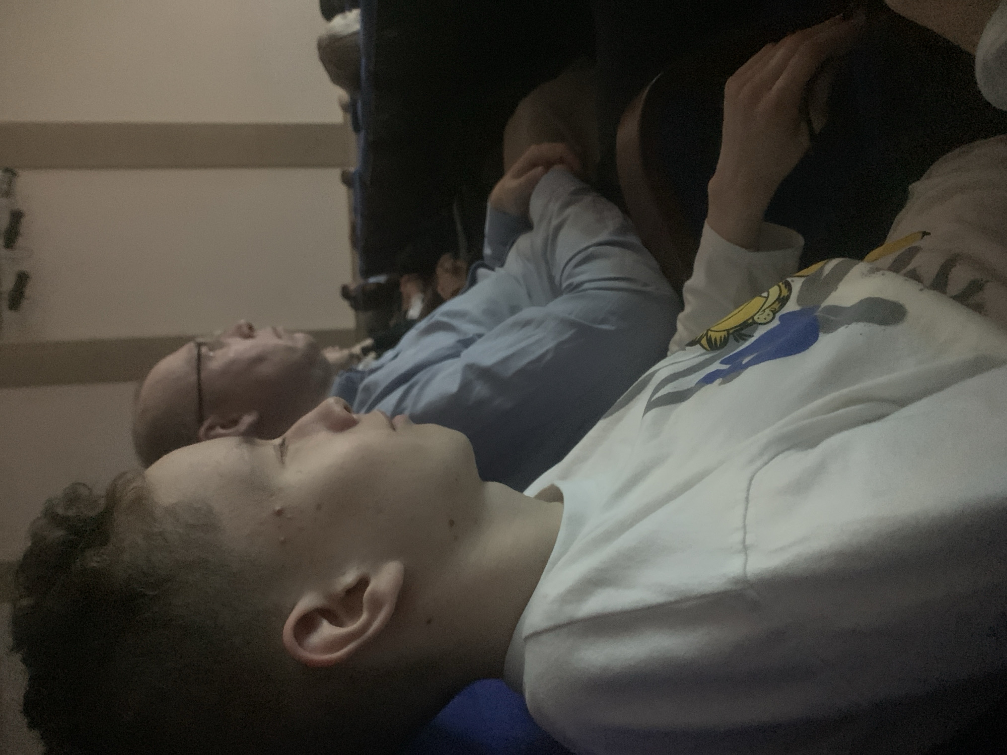 Two people are lying down. The person in the foreground is a young person with short, curly dark hair. They are wearing a white shirt. Behind them is an older bald man wearing glasses and a light blue shirt. They are lying in a dark room, possibly a movie theater. The location, Izhevsk, Russia, is unknown and not visible in the image.