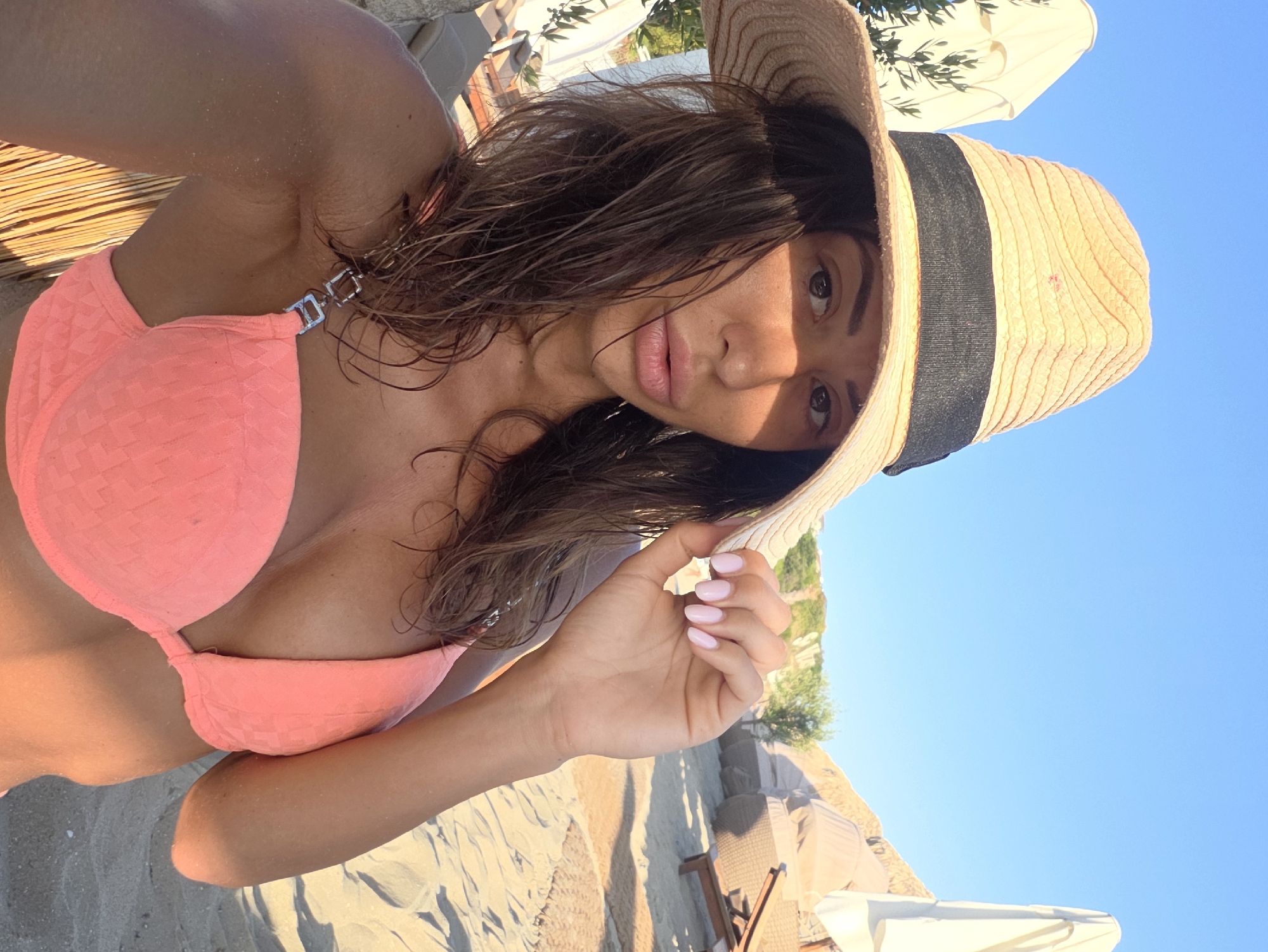 A woman wearing a straw hat and a pink bikini top poses for a selfie on a sandy beach. The woman has dark hair and is looking at the camera. The background shows beach umbrellas and lounge chairs. The scene appears to be outdoors on a sunny day. The location is identified as Sveti Vlas, Bulgaria.