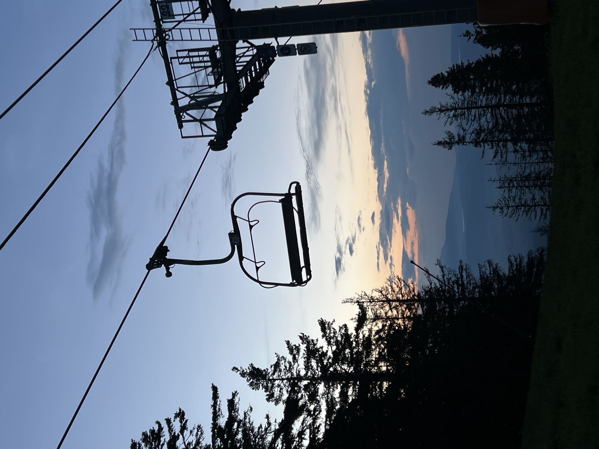 The image captures a ski lift chair suspended in silhouette against a dusky sky. The sky transitions from a pale blue at the top to hues of orange and pink near the horizon, with scattered clouds. Tall, dark silhouettes of pine trees frame the left side of the image, with a grassy slope in the foreground. A massive ski lift tower stands on the right, also in silhouette, with cables extending across the frame. The tower features ladders and signage. In the distance, layers of mountains are visible, receding into the haze. The scene evokes a sense of tranquility and the end of a day, possibly at the end of a ski season or the beginning of a new day. The ski lift and surrounding trees suggest a mountainous or forested resort area. While the image depicts a ski lift, it is not explicitly stated that this location is in the city of Samokov, Bulgaria.