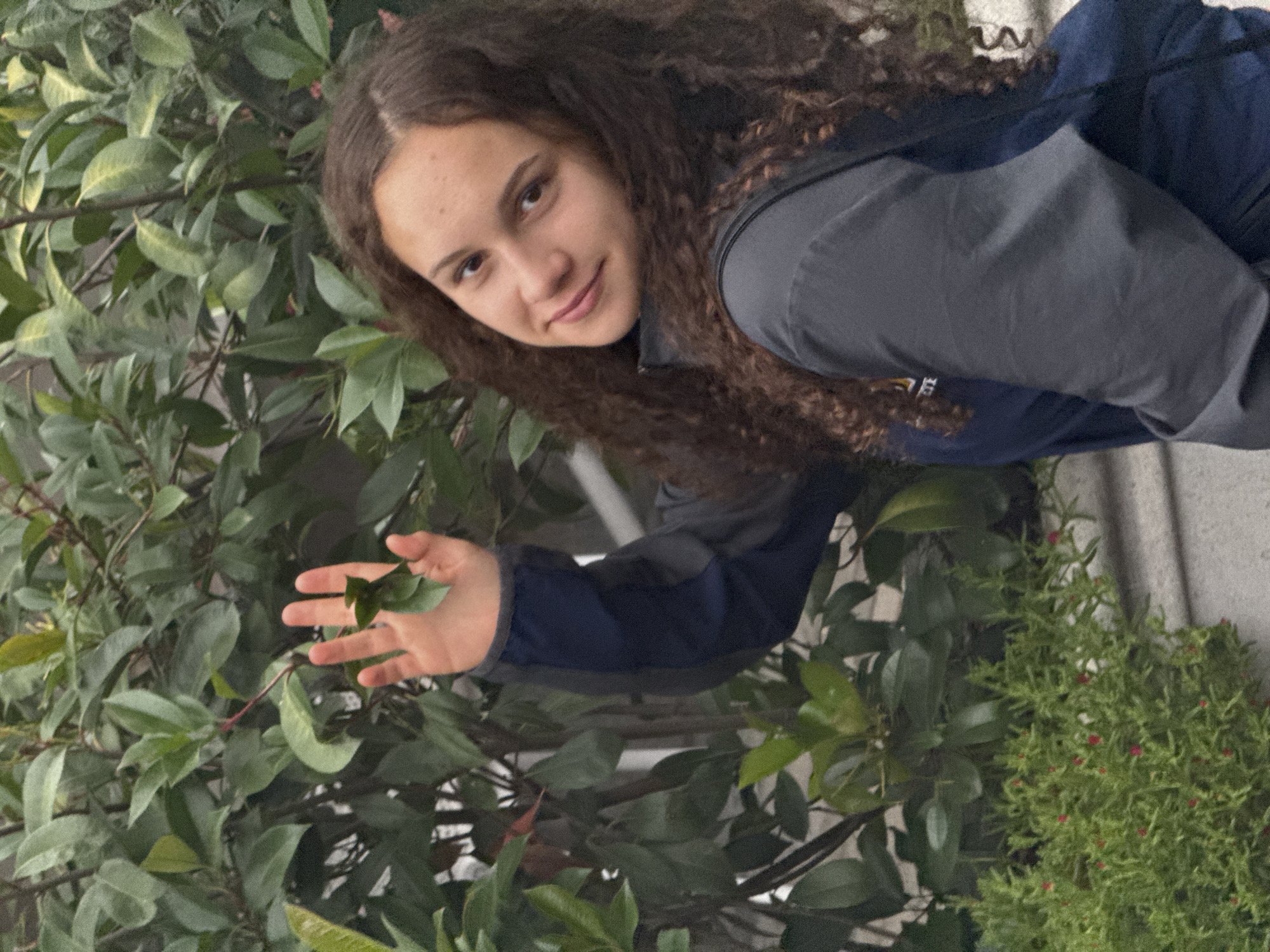 The image shows a young person with long, curly brown hair and fair skin, looking towards the camera with a slight smile. They are wearing a dark blue and gray jacket. Their left hand is reaching out to touch a green leaf on a plant with large, glossy leaves. In the background, there are more plants, including a bush with small red berries. The scene appears to be outdoors, possibly in a garden or near some greenery. The lighting suggests it could be daytime, though the exact time and weather are not discernible. The overall mood is casual and relaxed. There is no visible text in the image.