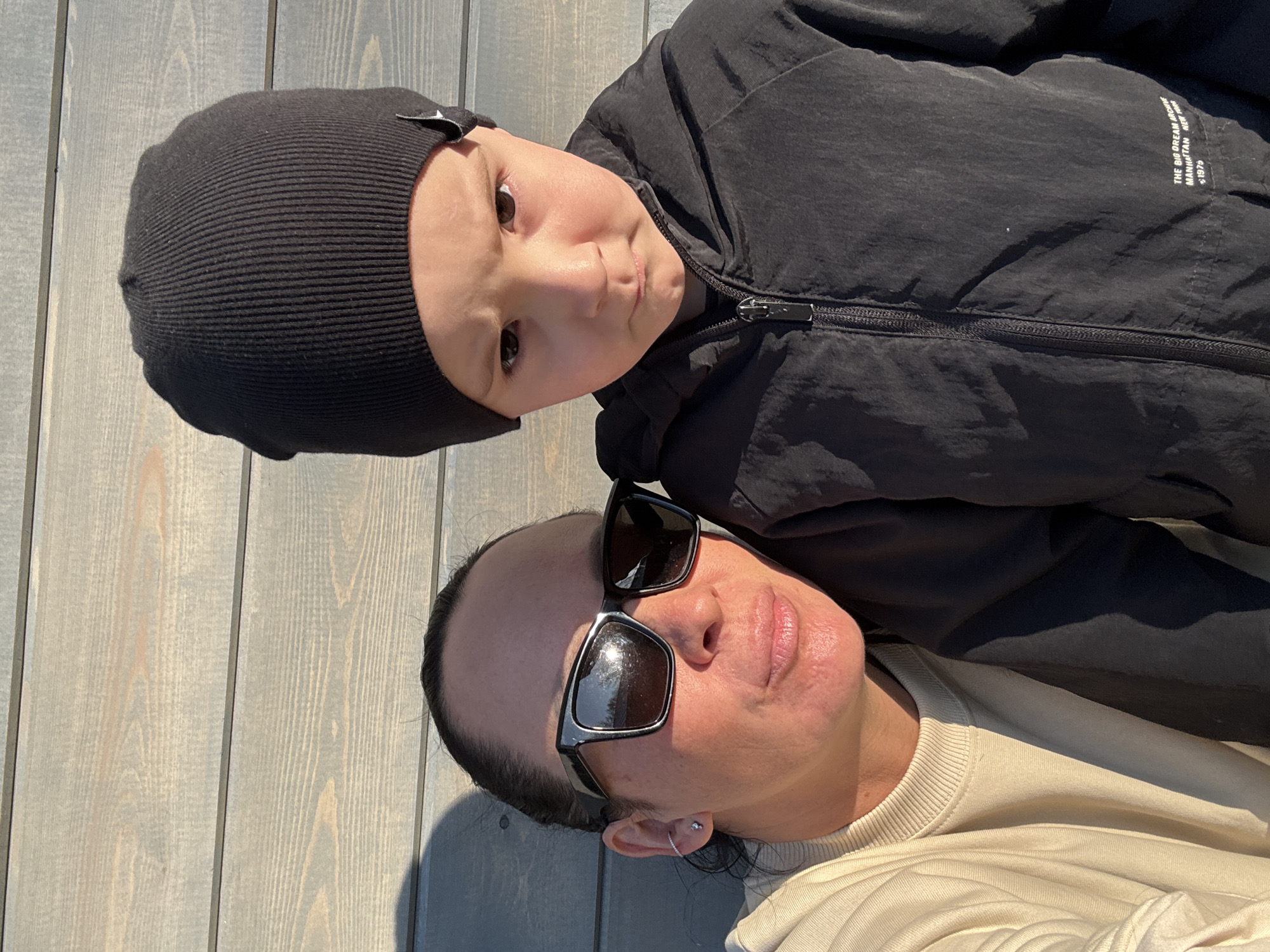 A selfie shows a fair-skinned adult and a fair-skinned child lying outdoors against a light gray, weathered wood surface. The adult wears sunglasses, a tan sweater, and an earring in their left ear. The child wears a black knit hat and a black, zippered jacket. The jacket has white text on its right side. The location is unknown and cannot be determined from the image.