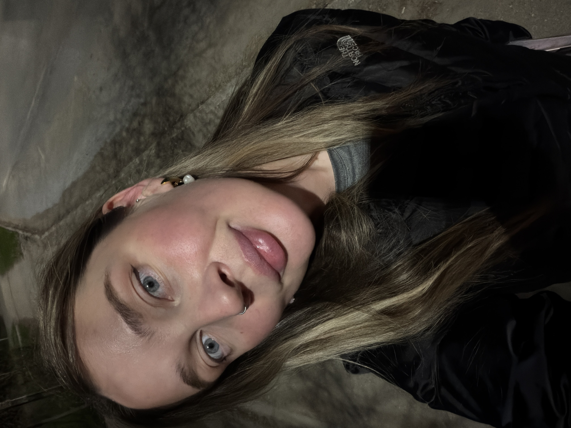 A woman with light blonde hair and blue eyes is pictured in a close-up, low-angle selfie. She is wearing a black jacket and a gray shirt underneath. She has a nose ring and earrings. The background appears to be an outdoor setting at night, potentially in a city. There is a blurred concrete surface visible, which could be a sidewalk or a road. The city of Chaikovsky, Russia, is not discernible in the image. The environment is dark with artificial lighting.