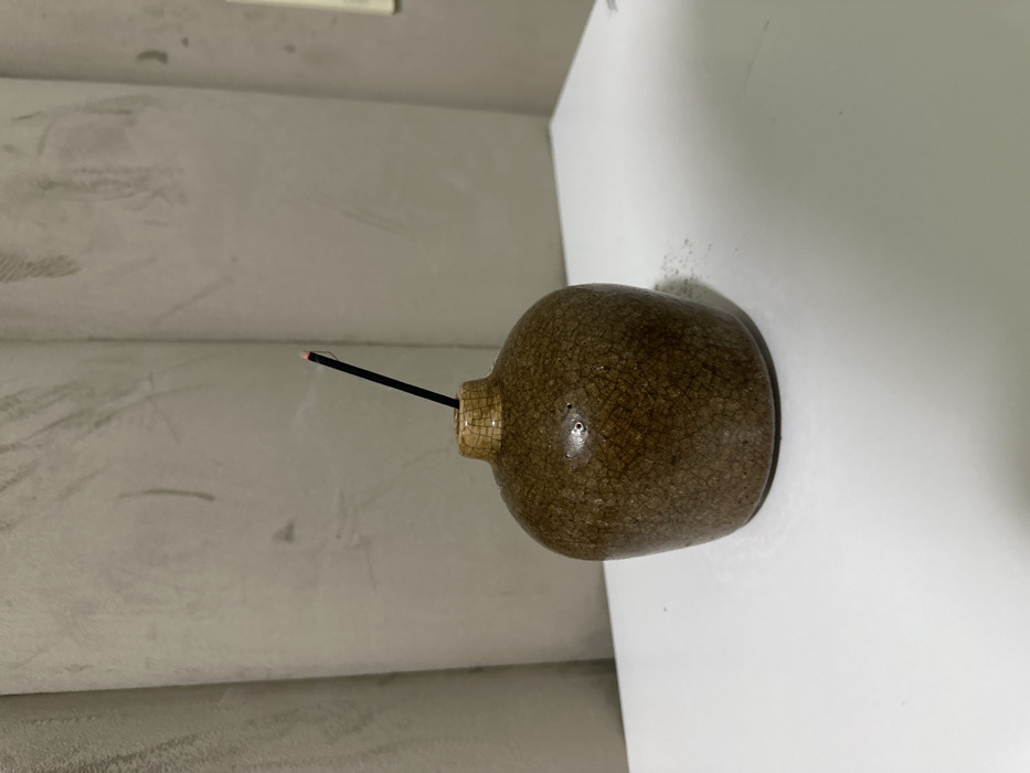 A brown, spherical ceramic incense holder with a visible crackle glaze rests on a clean white horizontal surface. A slender, black incense stick is vertically inserted into a narrow opening at the top of the holder. The tip of the incense stick glows red, and a faint wisp of smoke rises from it. The background consists of two light grey, textured wall panels or curtains meeting at an obtuse angle, indicating an indoor setting in Mexico City, Mexico. No other subjects or people are present.