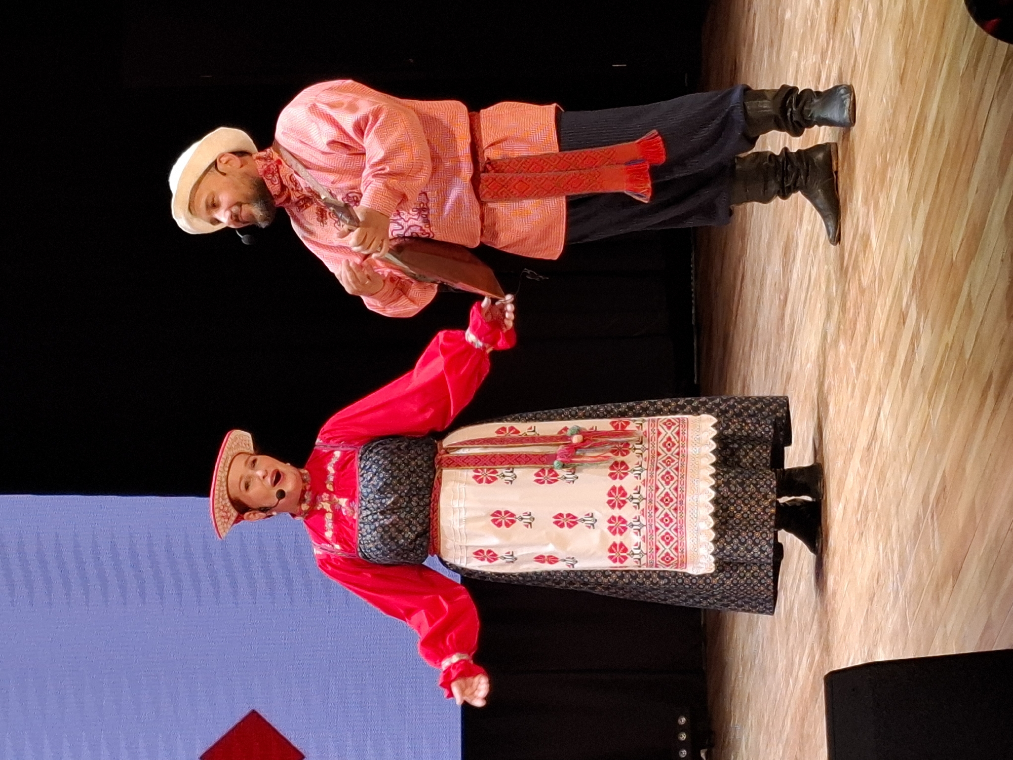 In a dimly lit theater, two performers, a woman and a man, stand on a wooden stage in front of a dark backdrop and a large screen displaying a light blue and red geometric pattern. The woman, on the left, wears a traditional red and white embroidered Russian folk costume. Her costume includes a red blouse with voluminous sleeves, a dark patterned skirt worn over a white apron adorned with red floral embroidery and intricate geometric patterns, and a tall, ornate headdress. She holds her arms out to the sides and has a microphone near her mouth, suggesting she is singing or speaking. The man, on the right, is dressed in a vibrant pink embroidered shirt, dark pants, and black boots. He wears a white cap and has a beard. He is playing a traditional stringed instrument, possibly a balalaika, and also has a microphone. The stage lighting illuminates the performers and the wooden floor. The scene likely depicts a cultural performance or concert. While the location is mentioned as Kungur, Russia, there are no specific visual cues in the image to confirm this detail.