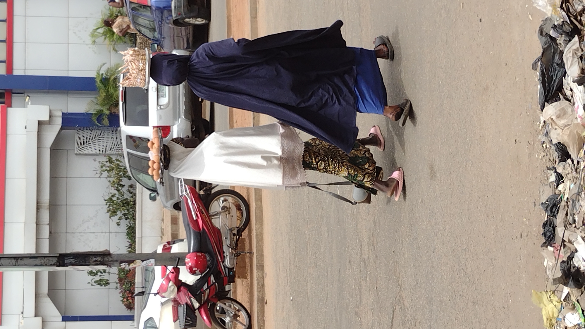 This daytime image captures a street scene in Jos, Nigeria, focusing on two women engaged in street vending. Both women are walking along an asphalt road, carrying goods on trays balanced on their heads.
The woman in the foreground wears a white long-sleeved garment over a patterned skirt and pink slide sandals. She carries a round, dark tray laden with numerous eggs on her head. Behind her, another woman is dressed in a dark blue long-sleeved garment over a blue skirt, paired with dark sandals. She also balances a round tray on her head, which holds a large quantity of what appear to be small, fried snacks or dough balls.
In the background, several vehicles are present, including a silver SUV and a partially visible blue sedan. A red motorcycle with a matching red helmet is parked near the women. Buildings with white and blue facades, some featuring red striped elements, line the left side of the street, interspersed with green foliage. A prominent detail on the right side of the image is a substantial pile of refuse, consisting of plastic bags and various discarded items, accumulated along the roadside. The lighting suggests it is a clear day. No discernible text is visible.