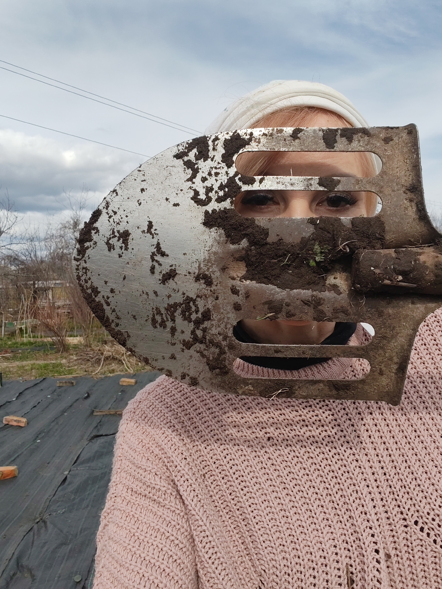 A woman with light blonde hair and a white head covering is holding a dirty spade in front of her face. Only her eyes and part of her mouth are visible through the holes in the spade. She is wearing a pink knitted sweater. The background shows a garden with black plastic covering the ground and bricks scattered around. In the distance, there are trees and a cloudy sky. The photograph was taken in the city of Chaikovsky, Russia.
