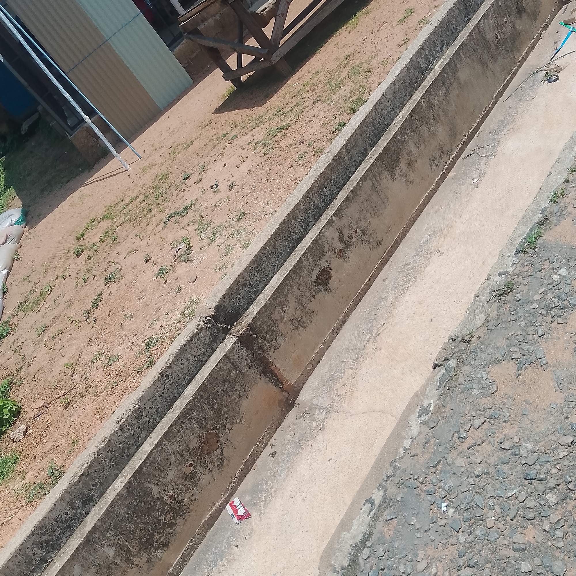 The image shows an outdoor scene in Jalingo, Nigeria. The main subjects are two parallel concrete drainage channels running diagonally across the frame. The ground beside the channels is composed of reddish-brown dirt with sparse green grass and some small pebbles. In the distance, there is a partially visible wooden structure and some corrugated metal sheeting, suggesting an industrial or construction area.
A small, folded piece of fabric with a red and white geometric pattern is visible lying in the bottom drainage channel, near the center of the image. There is also a blue rope hanging down from the top right edge of the frame, partially obscuring the concrete channel. The lighting suggests it is daytime, with shadows cast by the wooden structure. The weather appears to be dry, as the ground is dusty and there is no visible water in the channels. The overall impression is one of a functional, possibly under-maintained outdoor space. No people are visible in the image.