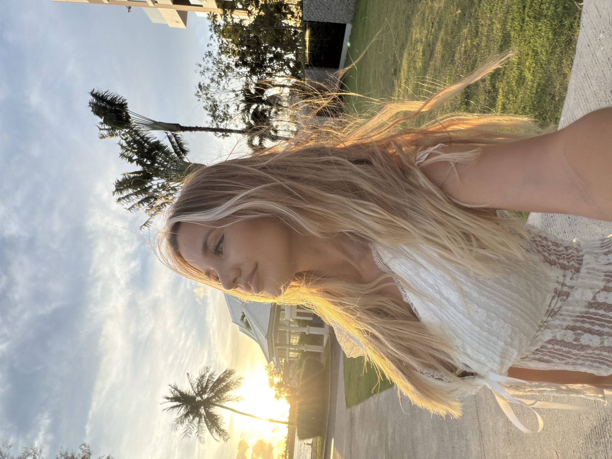 The media file captures a close-up, portrait-style selfie of a young woman with long, blonde hair. She is wearing a white lace top. The golden hour sunlight illuminates her hair and face, creating a warm and ethereal glow. Her eyes are looking down and to the side, with a soft, contemplative expression.
The background suggests a tropical setting, with palm trees visible and lush green grass. A modern building is partially visible on the left, and a white house with a pitched roof is seen in the distance, along with a body of water. The sky is partly cloudy, with streaks of light indicating the setting sun.
The overall mood is serene and peaceful, amplified by the soft lighting and the natural beauty of the surroundings. The image appears to have been taken outdoors in the late afternoon or early evening. There is no visible text in the media file. The location context is Choeng Thale, Thailand, which aligns with the tropical scenery.