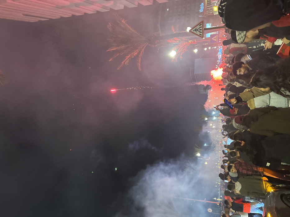 A nighttime scene shows a dense crowd gathered on a street, thick smoke filling the air. Red flares are prominent, adding to the haze. People are taking photos with their phones. Palm trees and streetlights are visible, along with signage. The crowd appears to be celebrating or protesting, judging by the atmosphere.